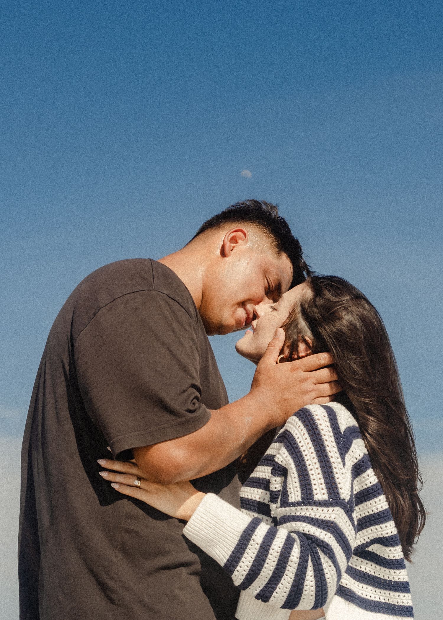 A couple sharing a tender moment outdoors under a clear blue sky, touching foreheads in a romantic embrace.