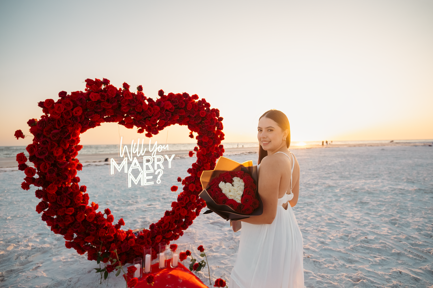 Woman in white dress smiles beside a red rose heart-shaped arrangement on a beach at sunset with Will You Marry Me text.