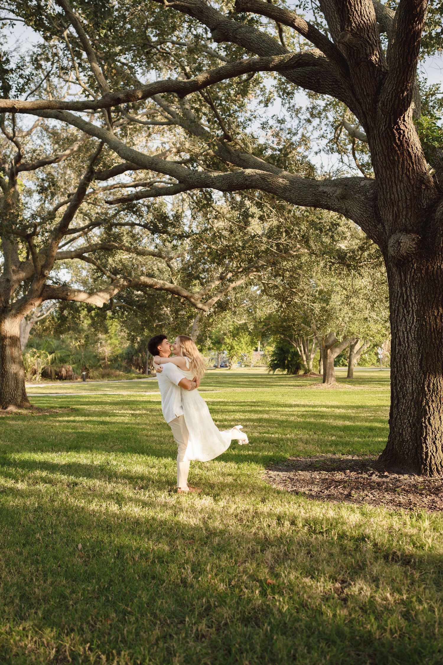 Couple in white attire embracing under large oak trees on lush green grass in a romantic outdoor setting.