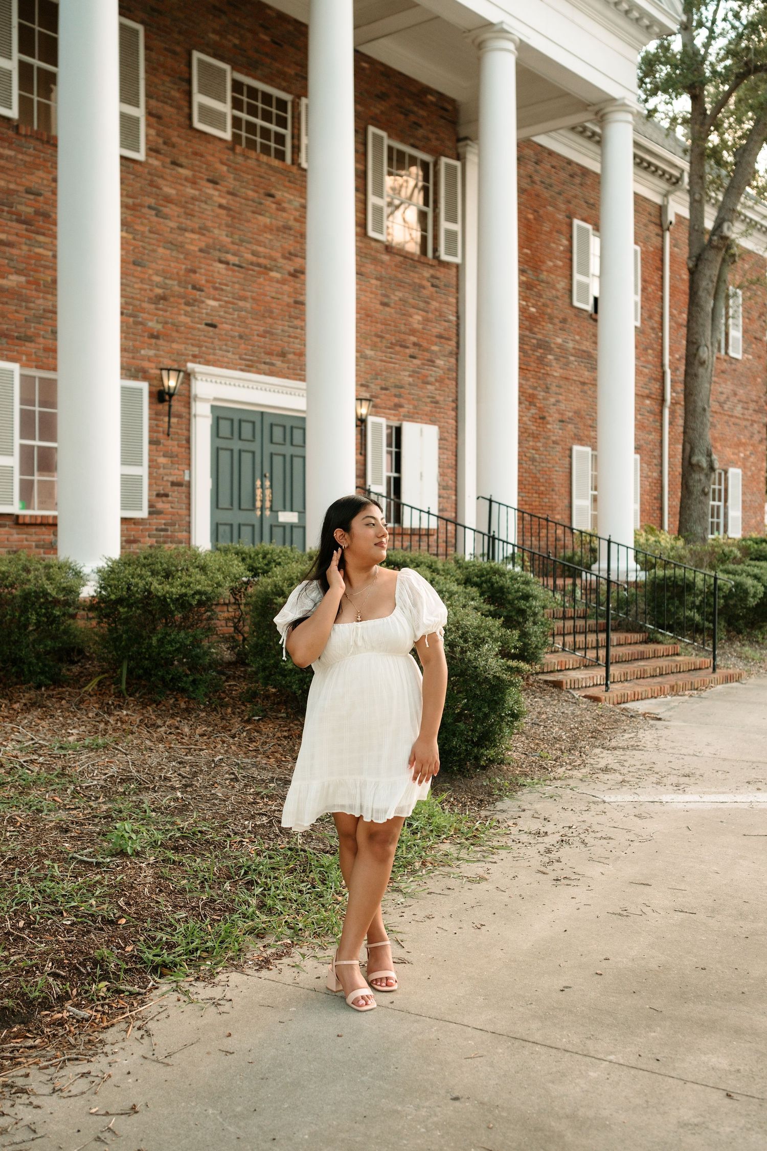 Woman in white mini dress posing outside a brick building with white columns and green door.