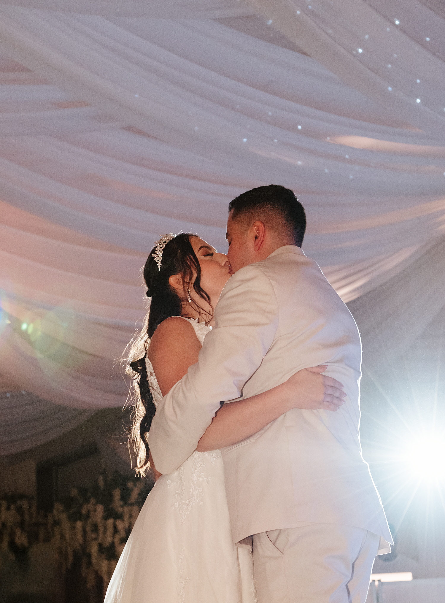Bride and groom share first kiss under elegant white draped ceiling at wedding reception.
