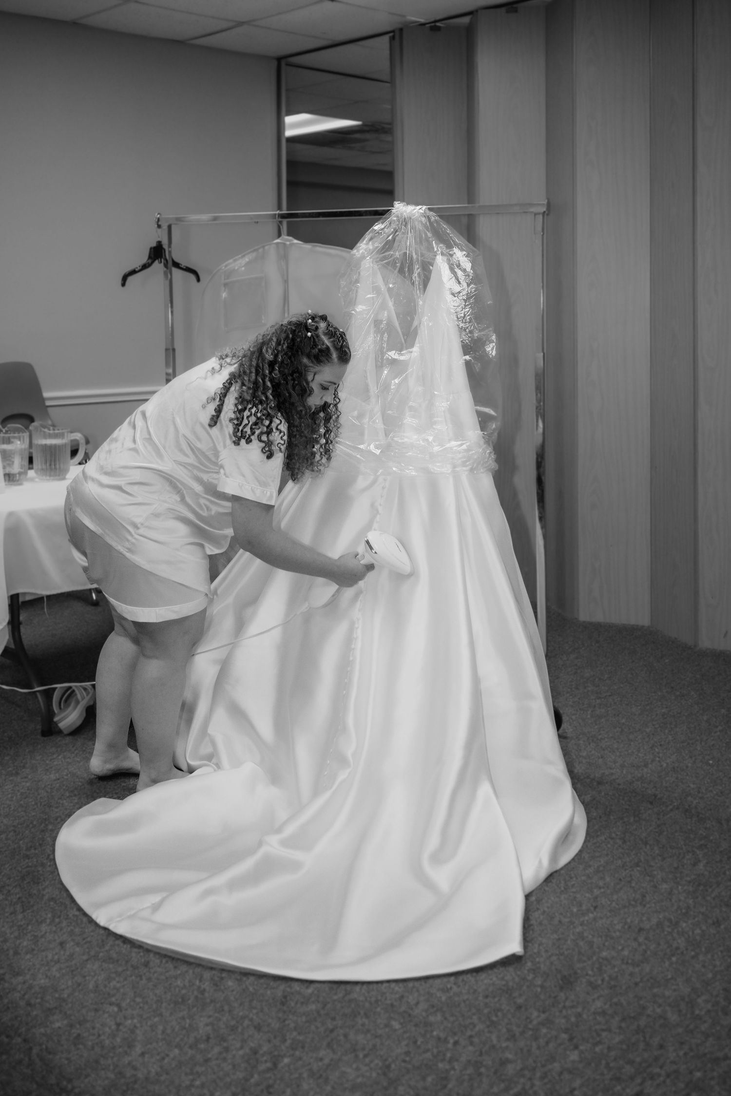 Black and white photo of a woman helping a bride adjust her wedding gown train in a bridal dressing room.
