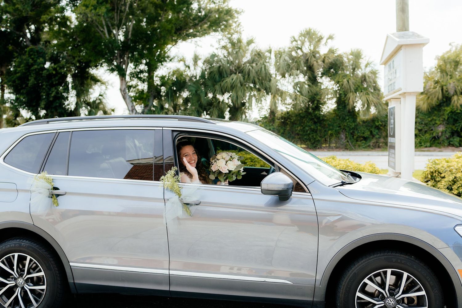 A bride sits in a silver SUV holding a white bouquet, decorated with white floral accents.