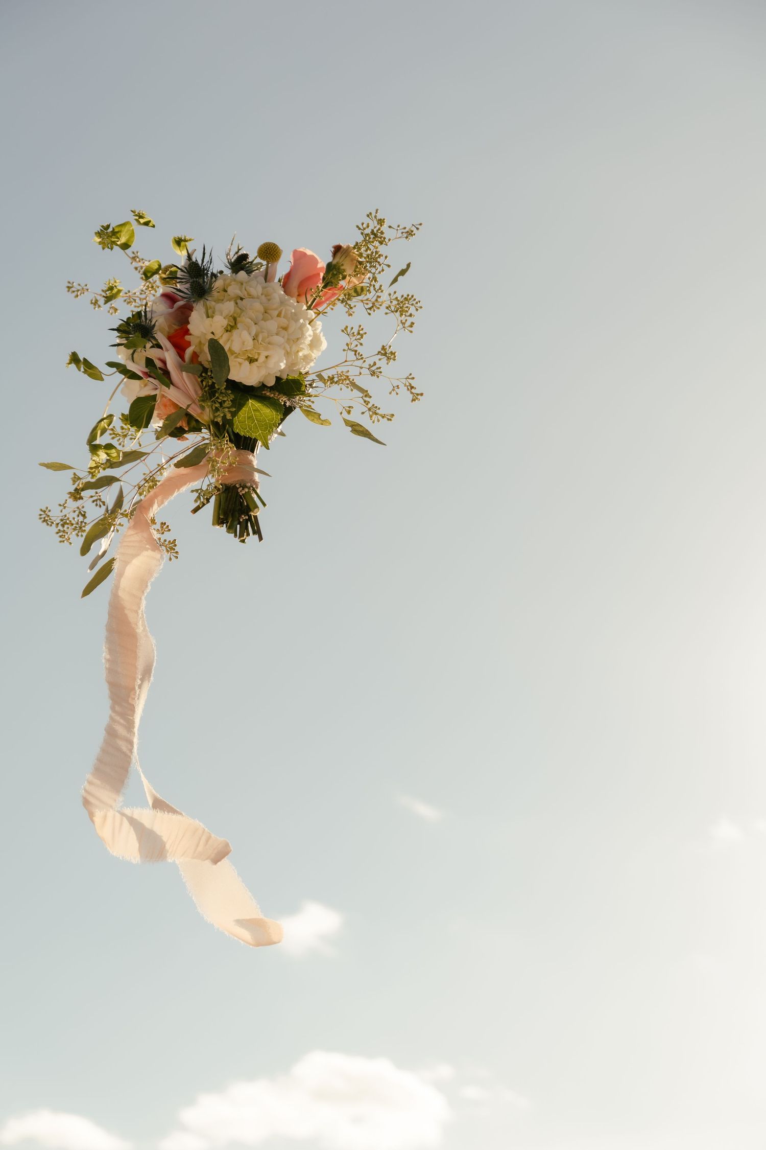 A bridal bouquet of white, pink flowers and greenery with flowing ribbons tossed against a clear blue sky.