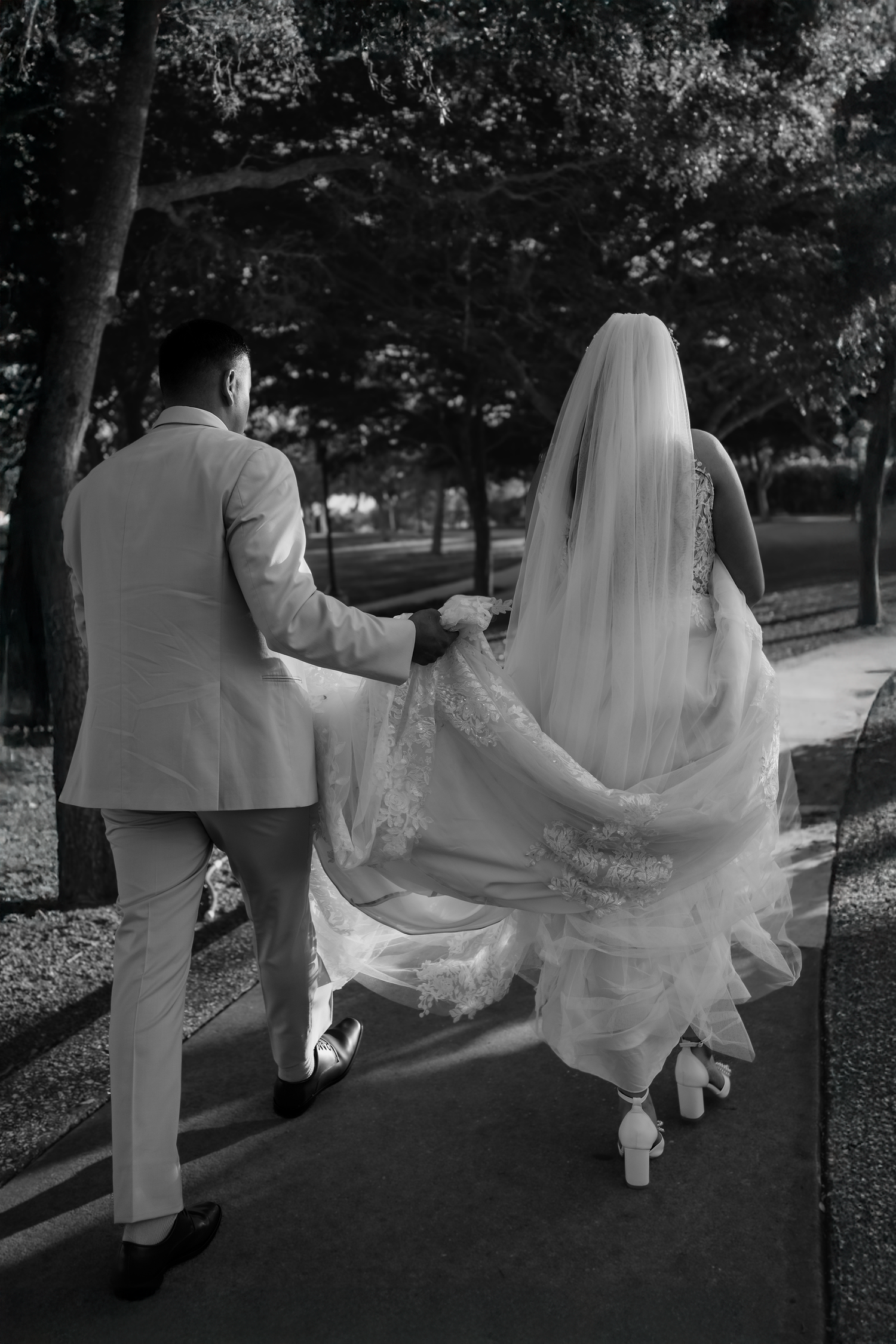 Black and white photo of bride and groom walking hand in hand along tree-lined path, wedding veil flowing behind.