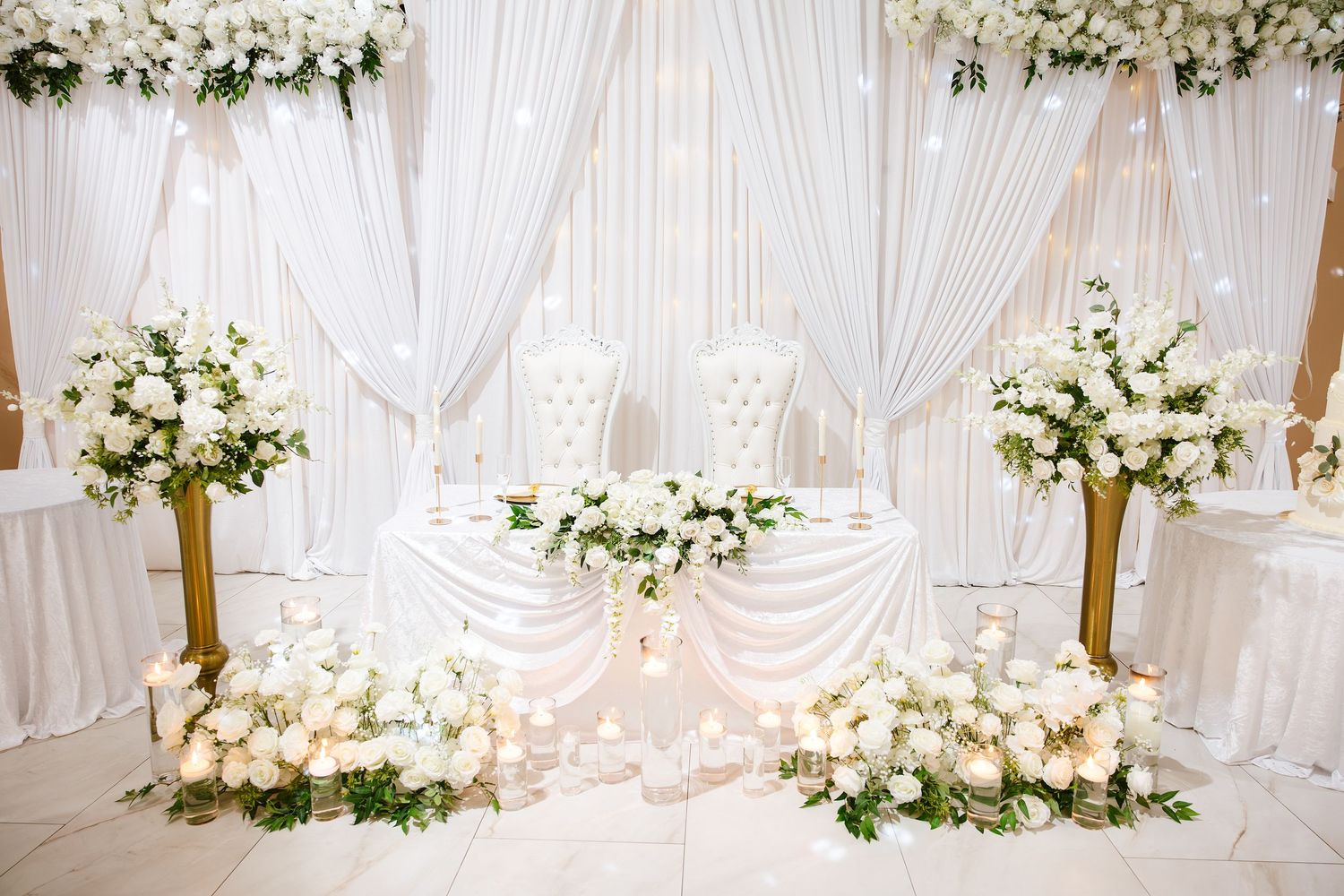 Elegant white wedding sweetheart table adorned with lush floral arrangements, candles, and draped fabric backdrop.