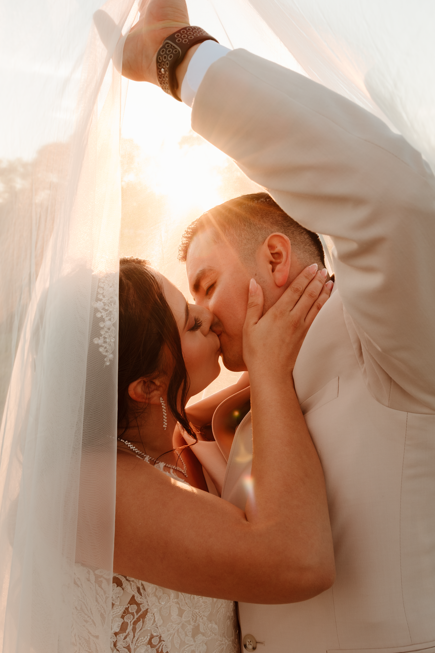 Romantic golden hour wedding photo of couple kissing under bride's flowing veil in warm sunlight.