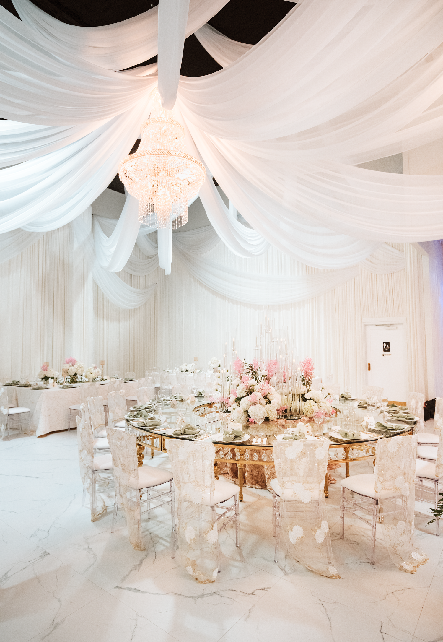 Elegant wedding reception with white draped ceiling, crystal chandelier, round glass tables, ghost chairs, and pink floral centerpieces.