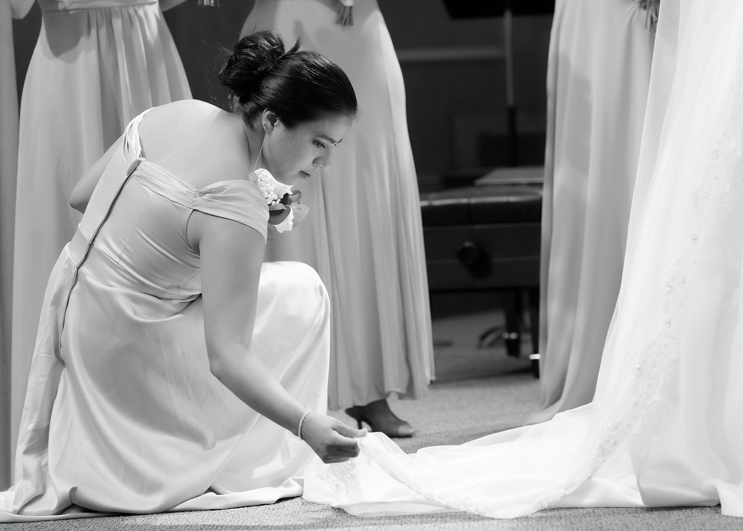 A woman in a white dress carefully adjusts the hem of a bridal gown in a black and white photo.