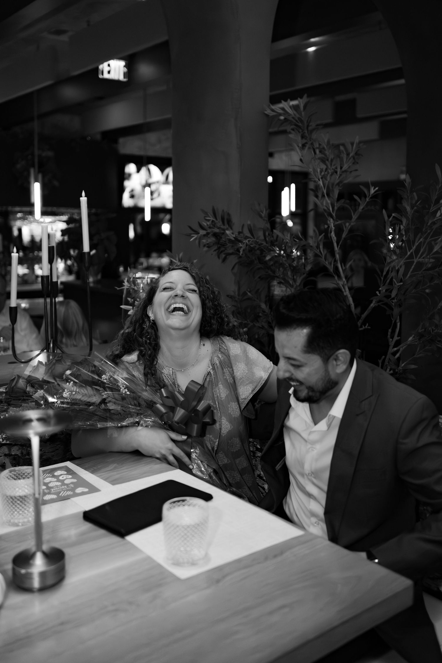 Couple laughing together at restaurant table with flowers and candles in black and white photo.