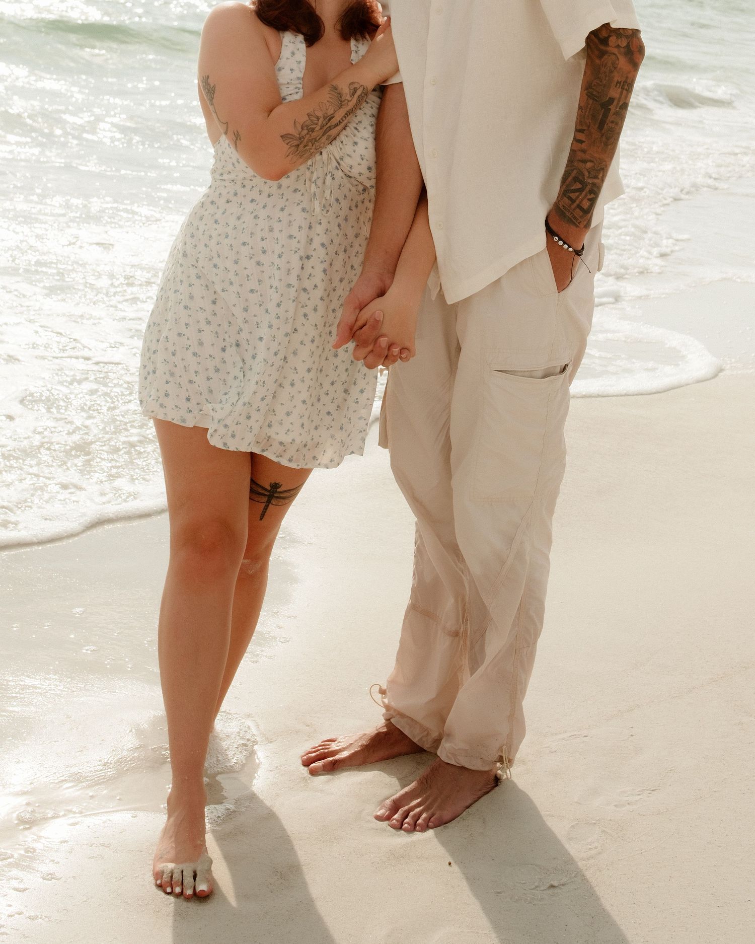 Couple with tattoos holding hands barefoot on a sandy beach, wearing white floral dress and linen pants near ocean waves.