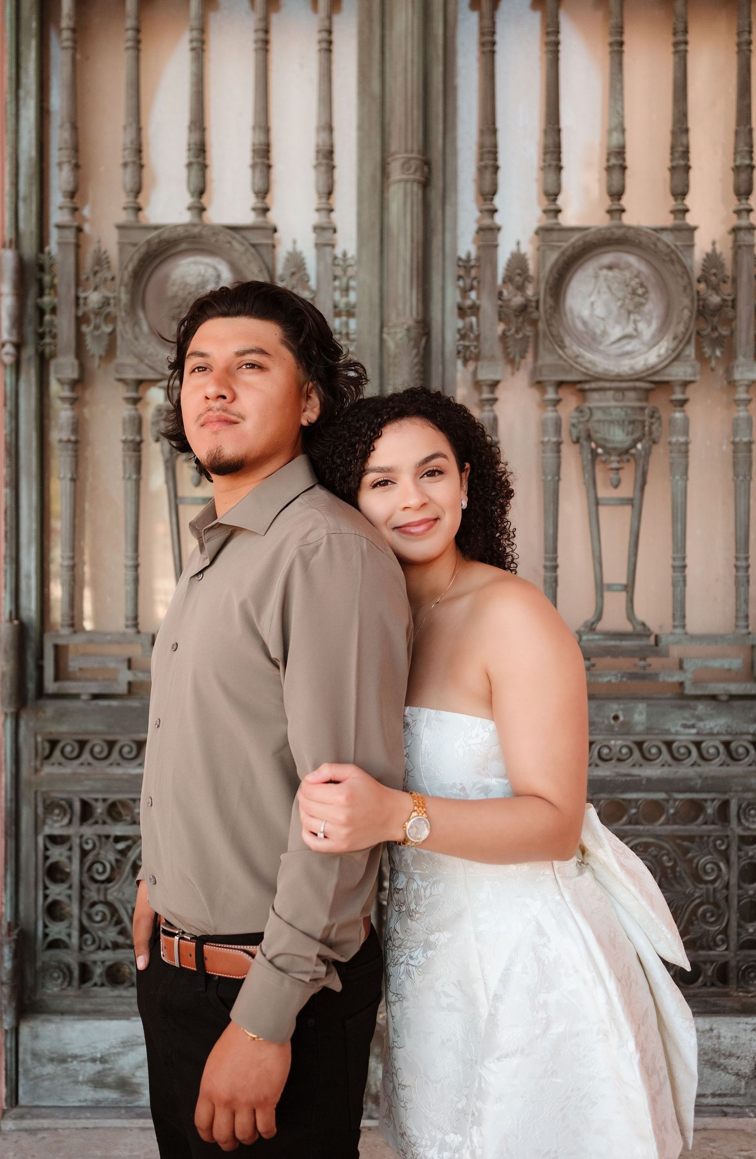 Couple posing in front of ornate bronze doors, woman in strapless white gown and man in tan shirt.