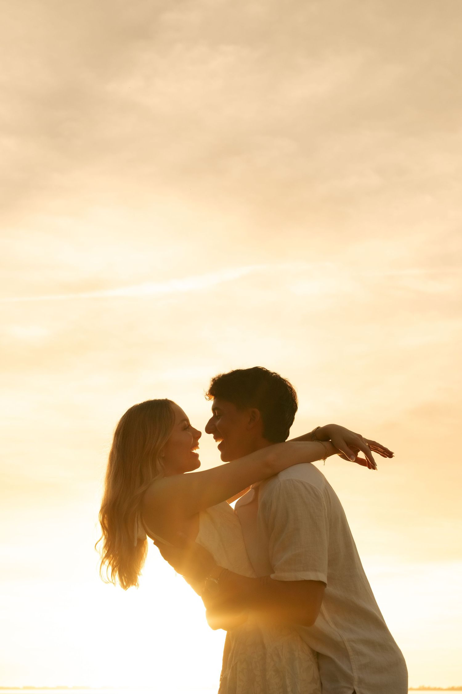 Romantic couple embracing at golden sunset, silhouetted against warm glowing sky outdoors.