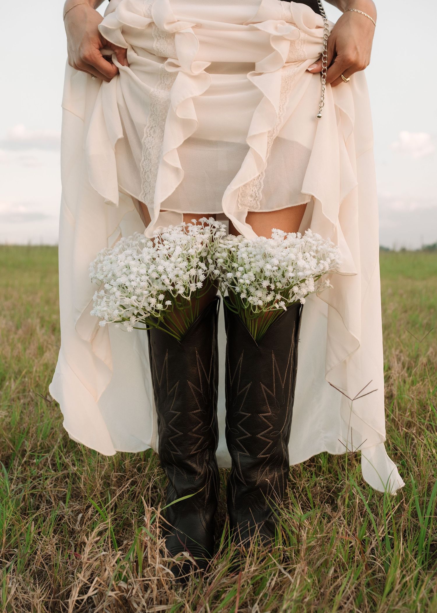 Bride in white ruffled dress and black cowboy boots holding baby's breath bouquet in a green field.