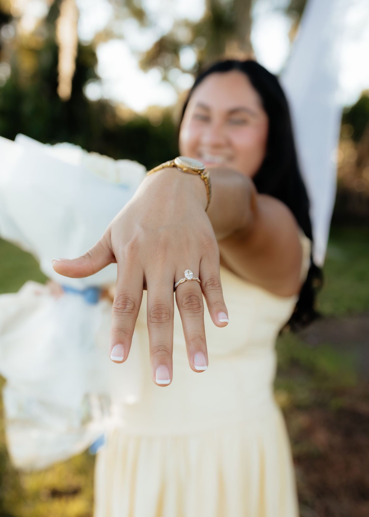 Woman in yellow dress smiling and showing off her engagement ring with a diamond solitaire outdoors.