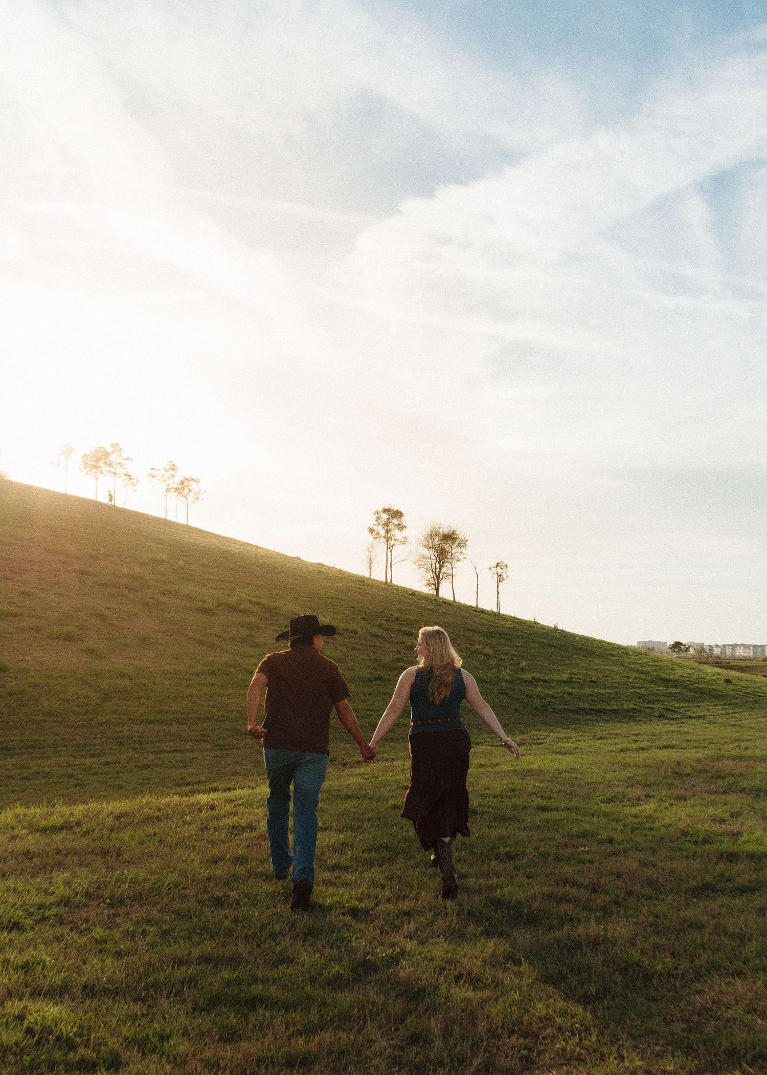 Couple holding hands walking up a sunlit green hill at golden hour, man wearing cowboy hat and woman in flowing dress.