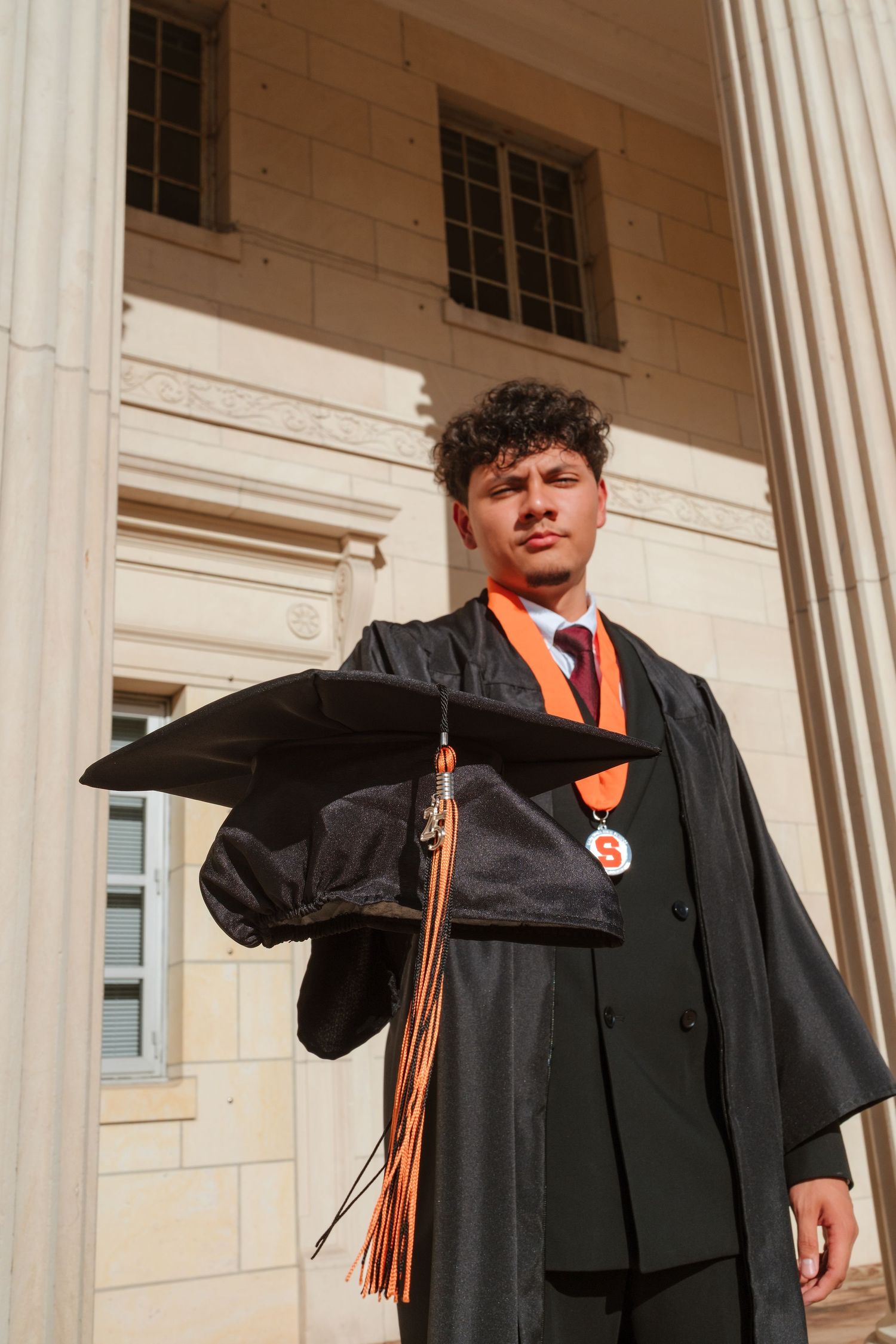 Graduate in black cap and gown holds mortarboard with orange tassel outside columned university building.