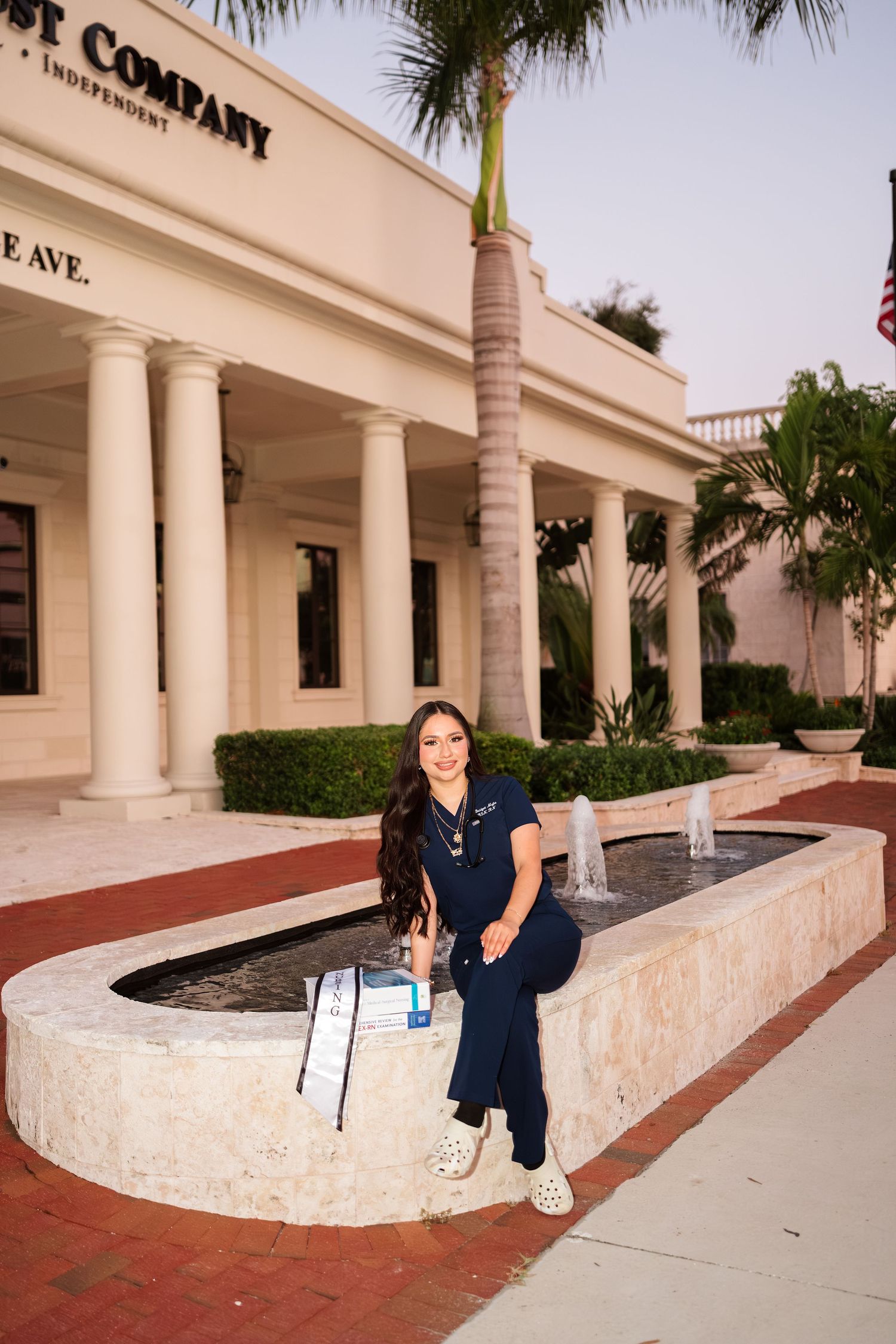 Woman in navy scrubs sitting on a fountain outside a trust company building with palm trees and columns.