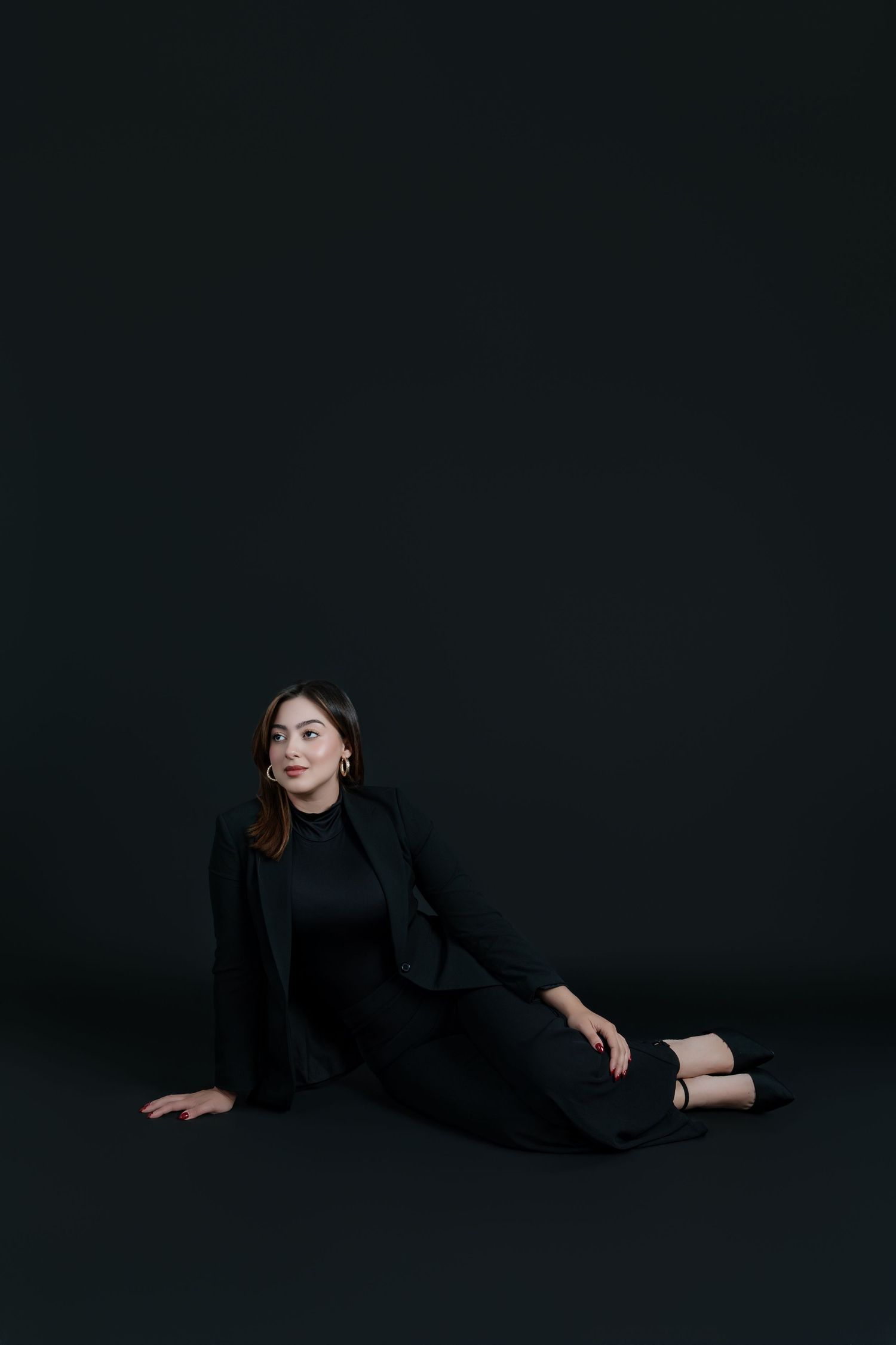 Elegant woman in black suit sitting on floor looking away against dark studio background.