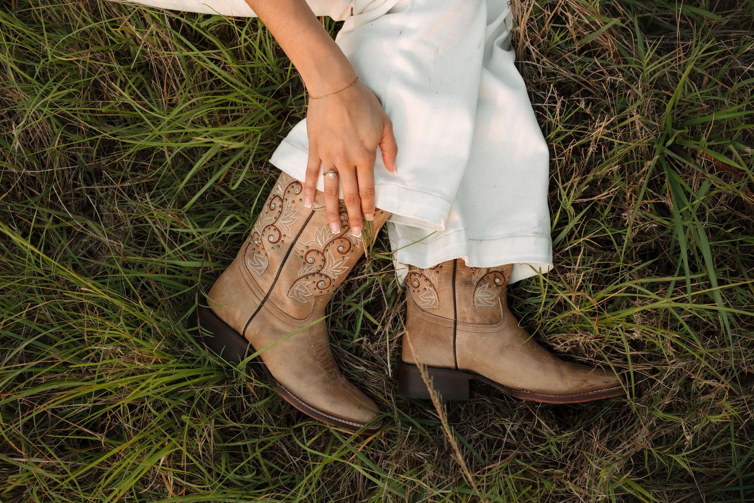 Woman in white pants and tan embroidered cowboy boots sitting in tall green grass, with her hand resting on her knee.
