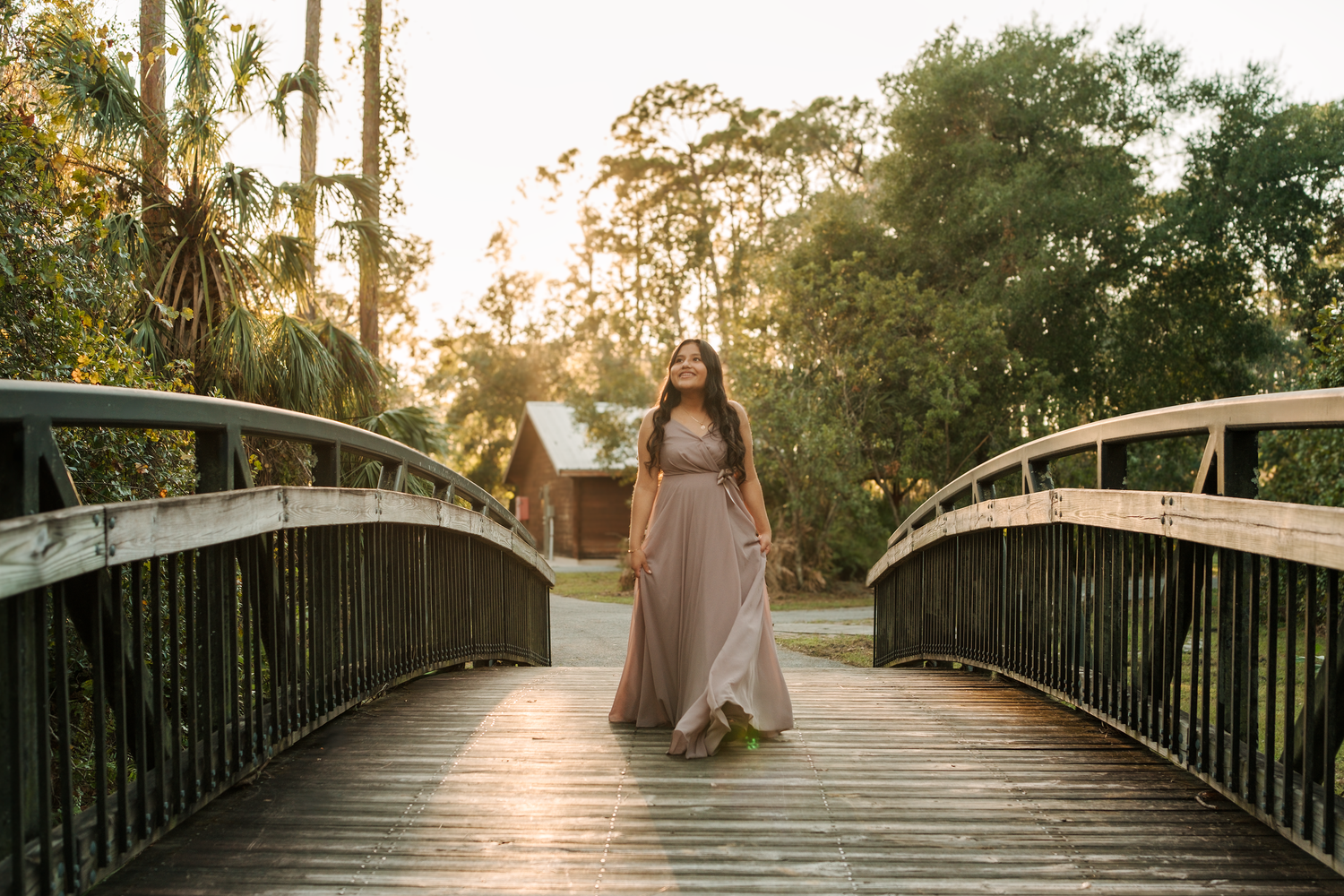 Woman in a long flowing dress walking barefoot across a curved wooden bridge at golden hour, surrounded by lush green trees and palms.