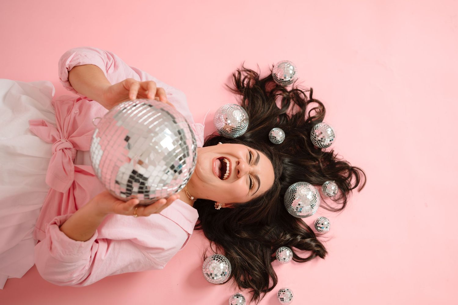 Woman in a pink outfit lies on a pink background, laughing while holding a large silver disco ball surrounded by small mirrored ornaments.