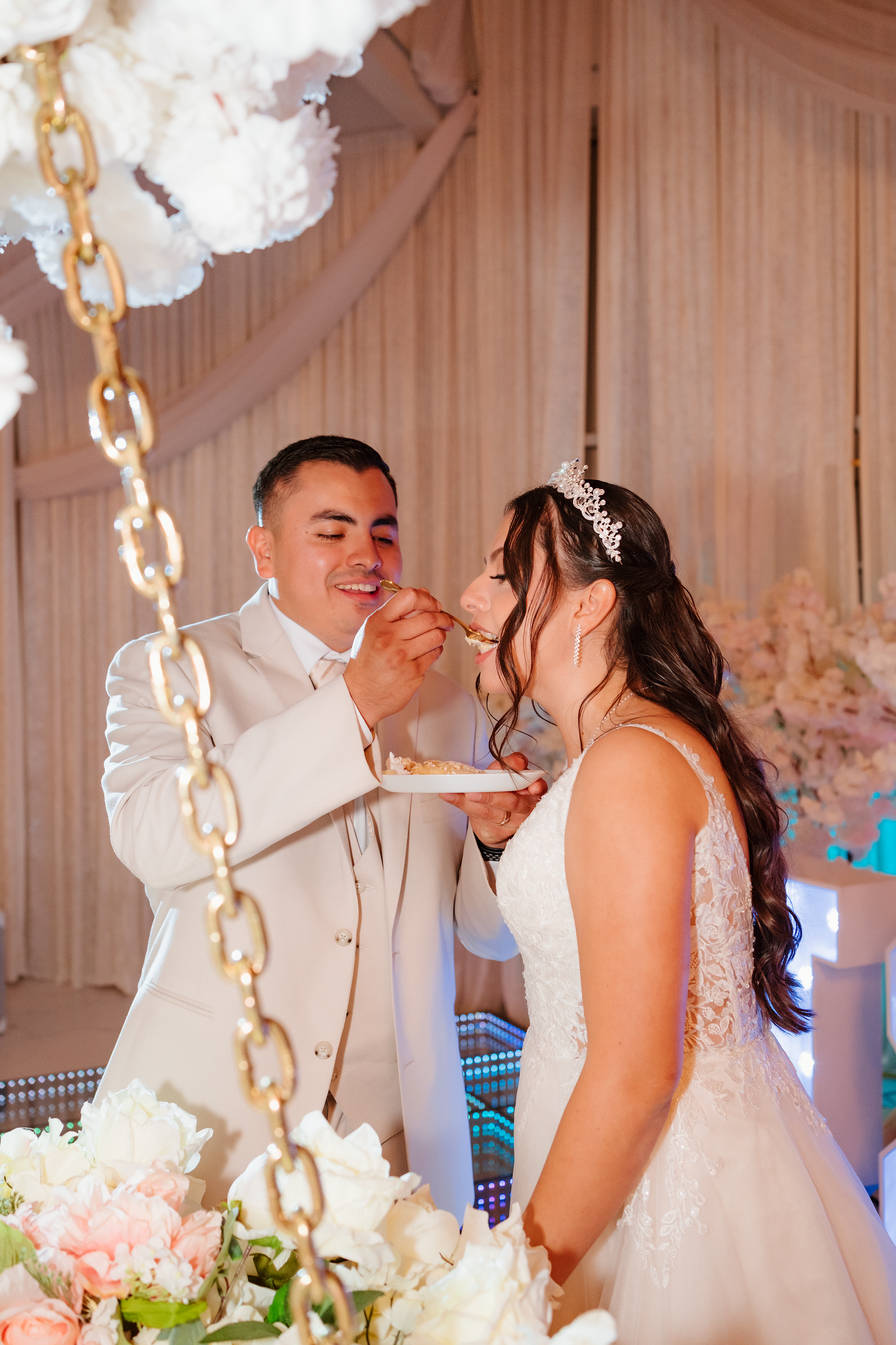 Smiling groom feeds bride wedding cake at elegant reception with floral decorations.