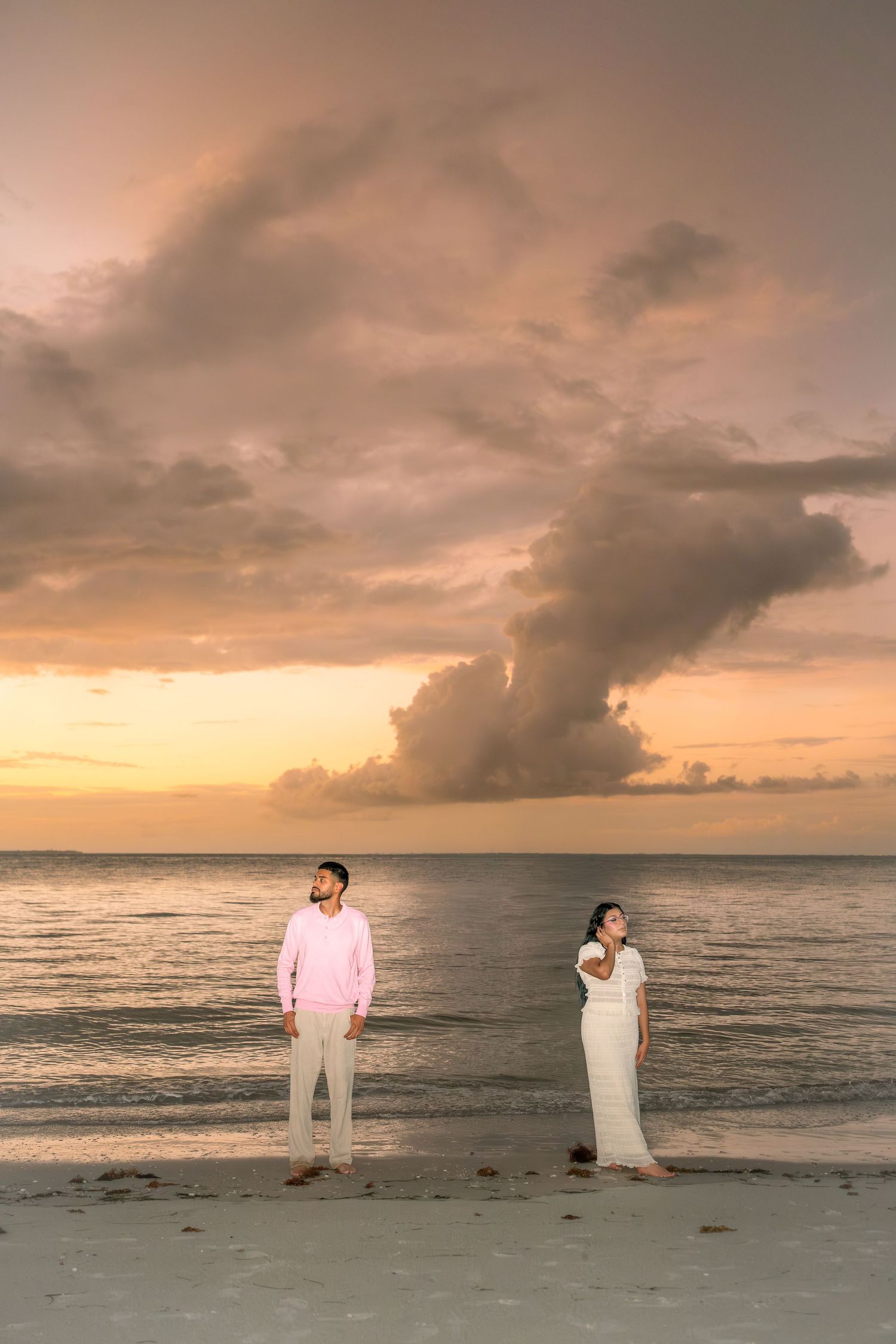 Couple standing apart on a beach at sunset, man in pink shirt, woman in white dress, dramatic cloudy sky.