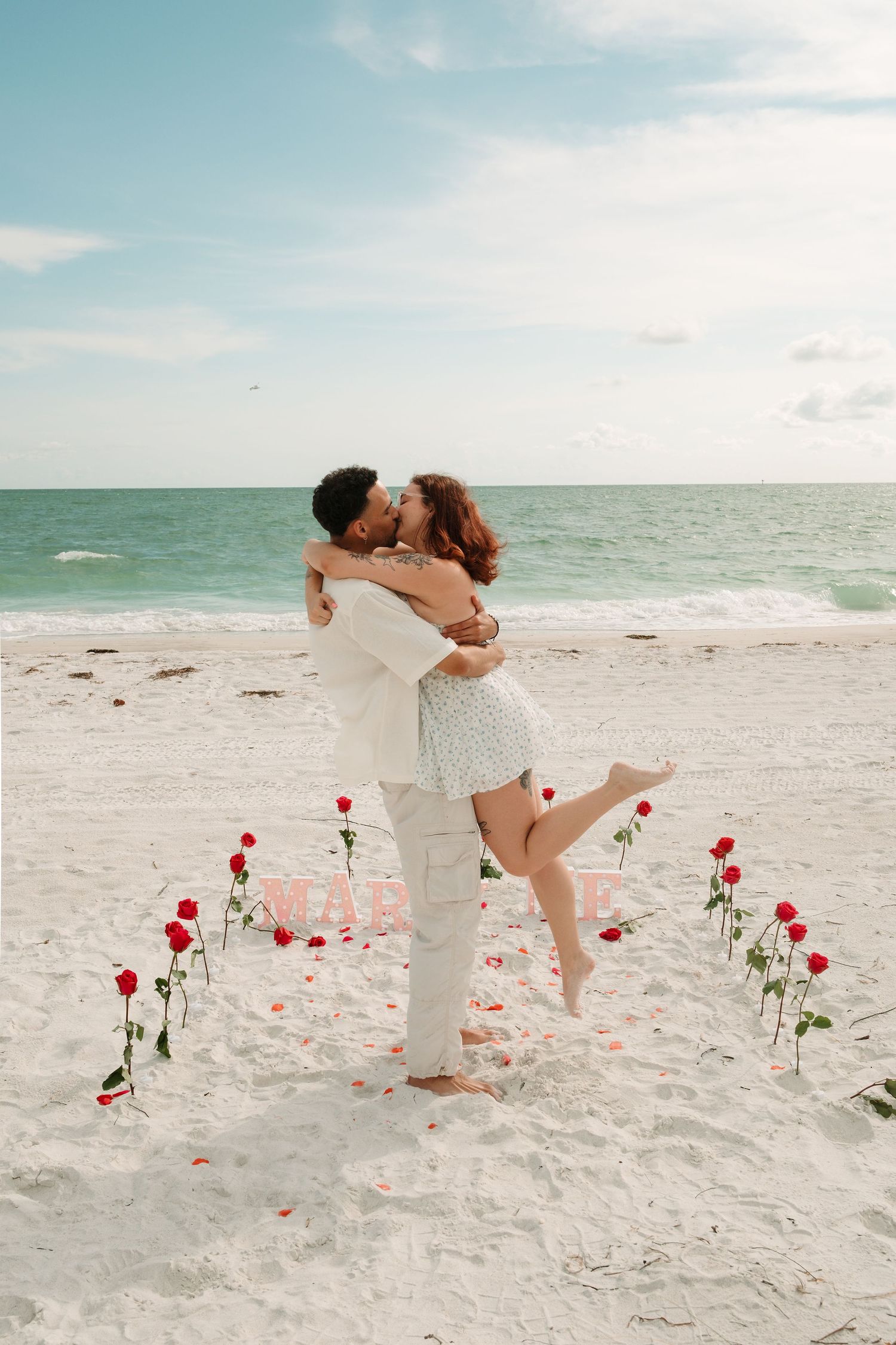 Couple kissing on a white sand beach with red roses arranged in a heart shape and a romantic marriage proposal setup.