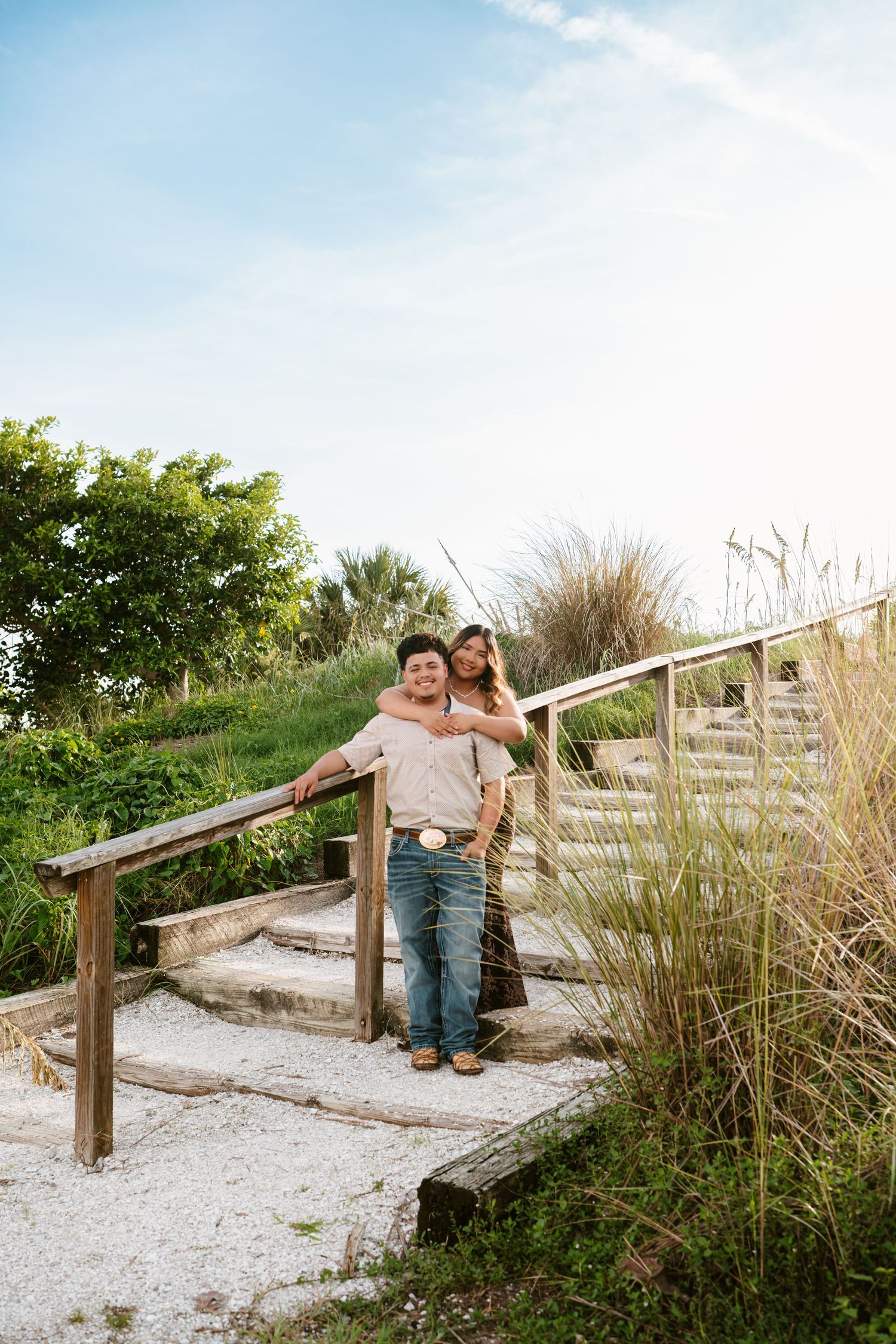 Couple embracing on rustic wooden steps surrounded by lush greenery and tall grass under a bright blue sky.