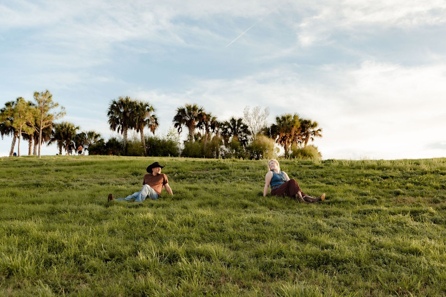 Two people relaxing on a lush green hillside surrounded by tall palm trees under a blue sky.