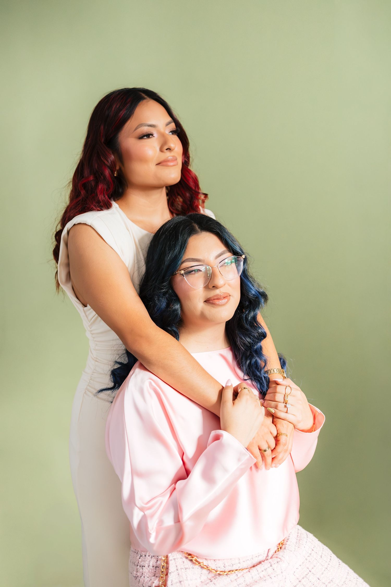 Two women posing together against a green background, one in white dress with red hair, other in pink with blue hair.