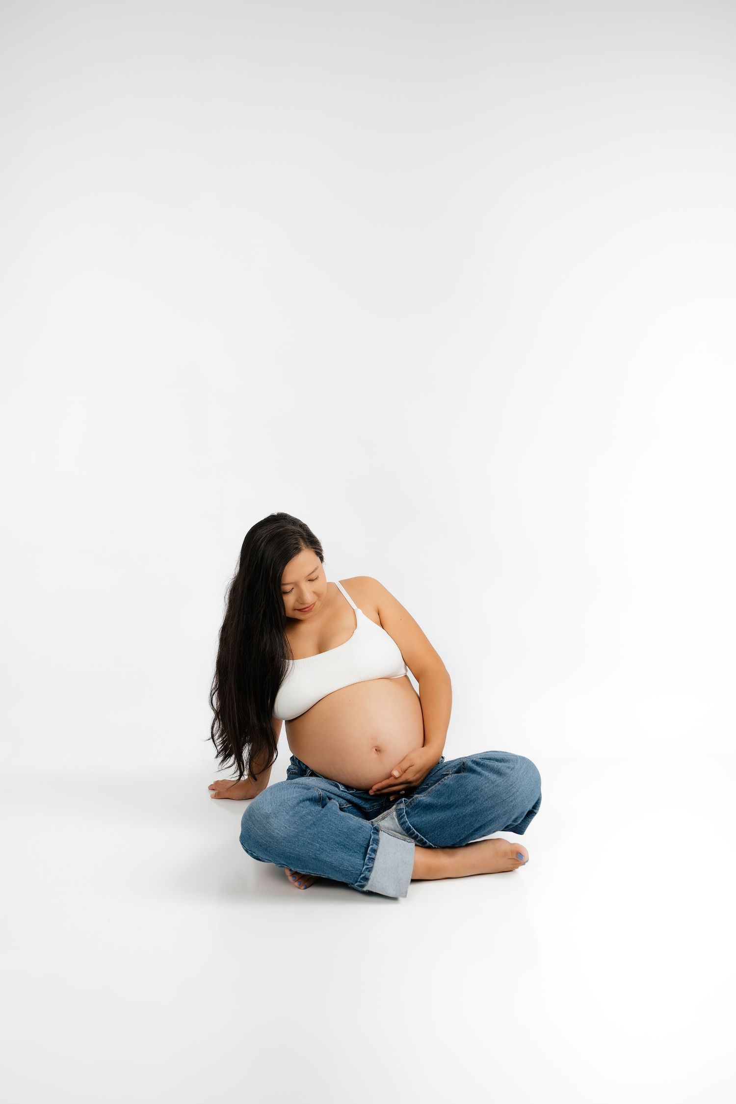 Pregnant woman in white crop top and blue jeans sitting cross-legged on white background, cradling her baby bump.
