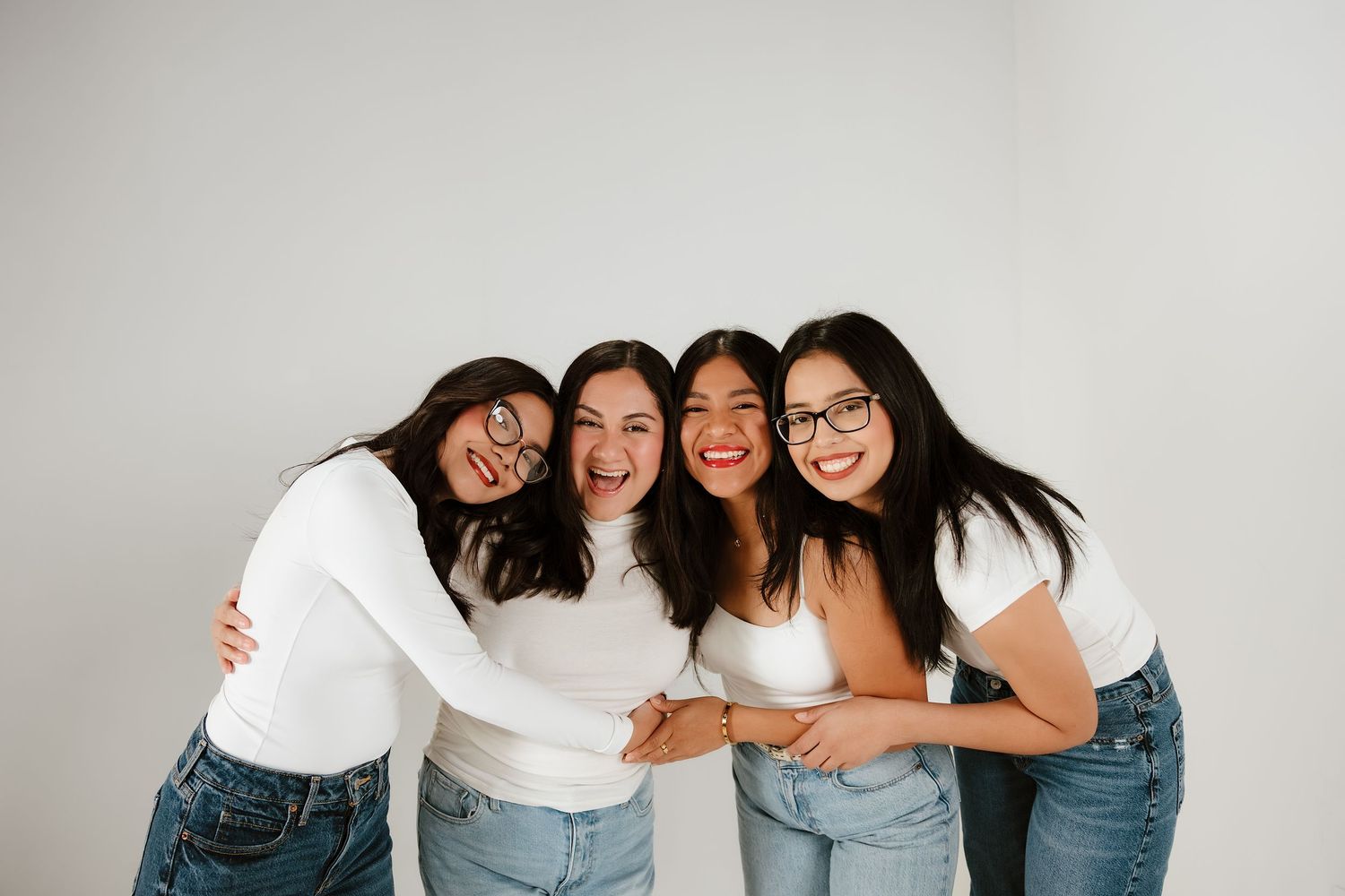 Four young women in white tops and jeans smiling and hugging together against a white background.