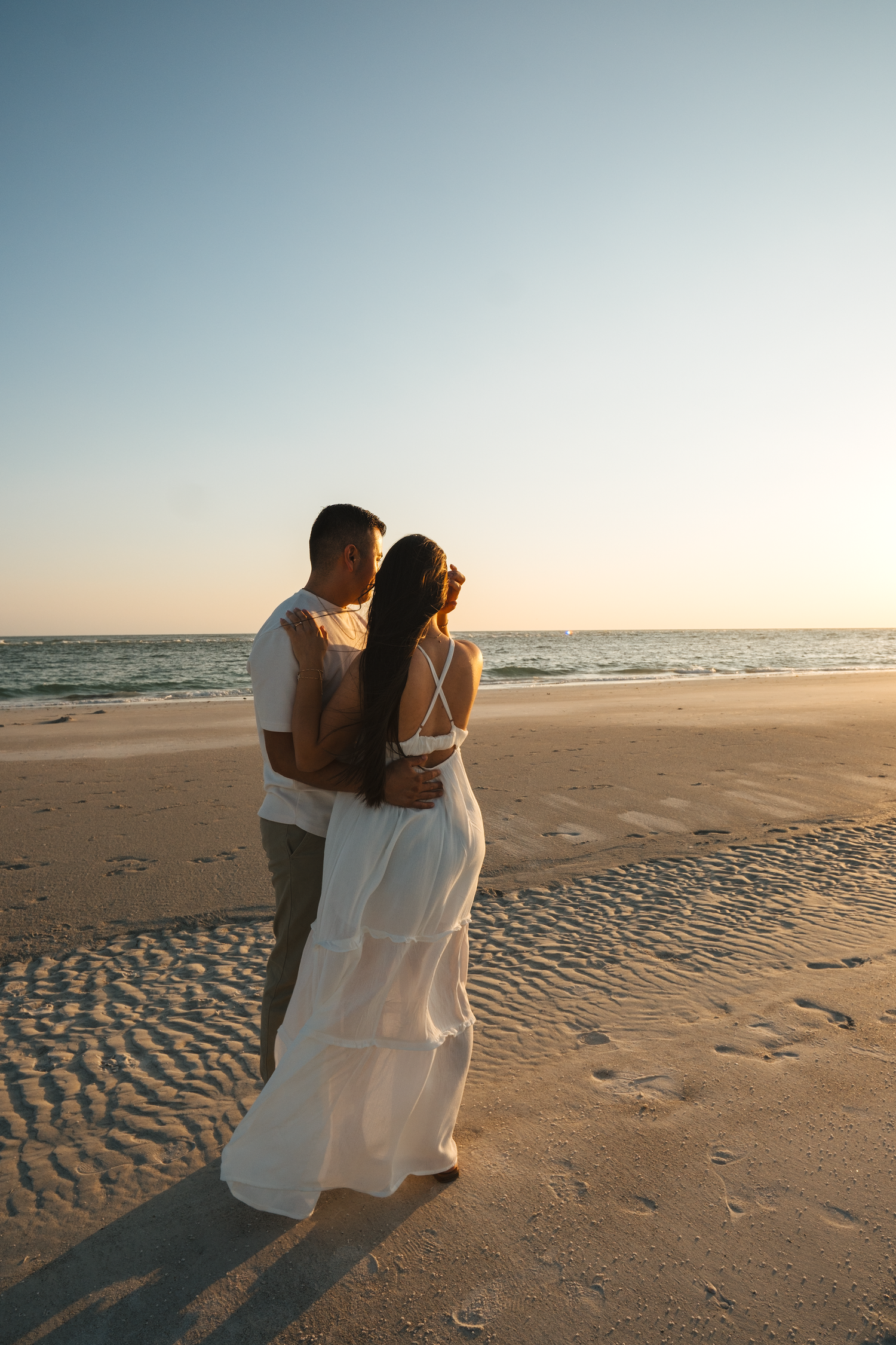 Couple in white embracing on a sandy beach at sunset, facing the calm ocean with footprints in the sand.