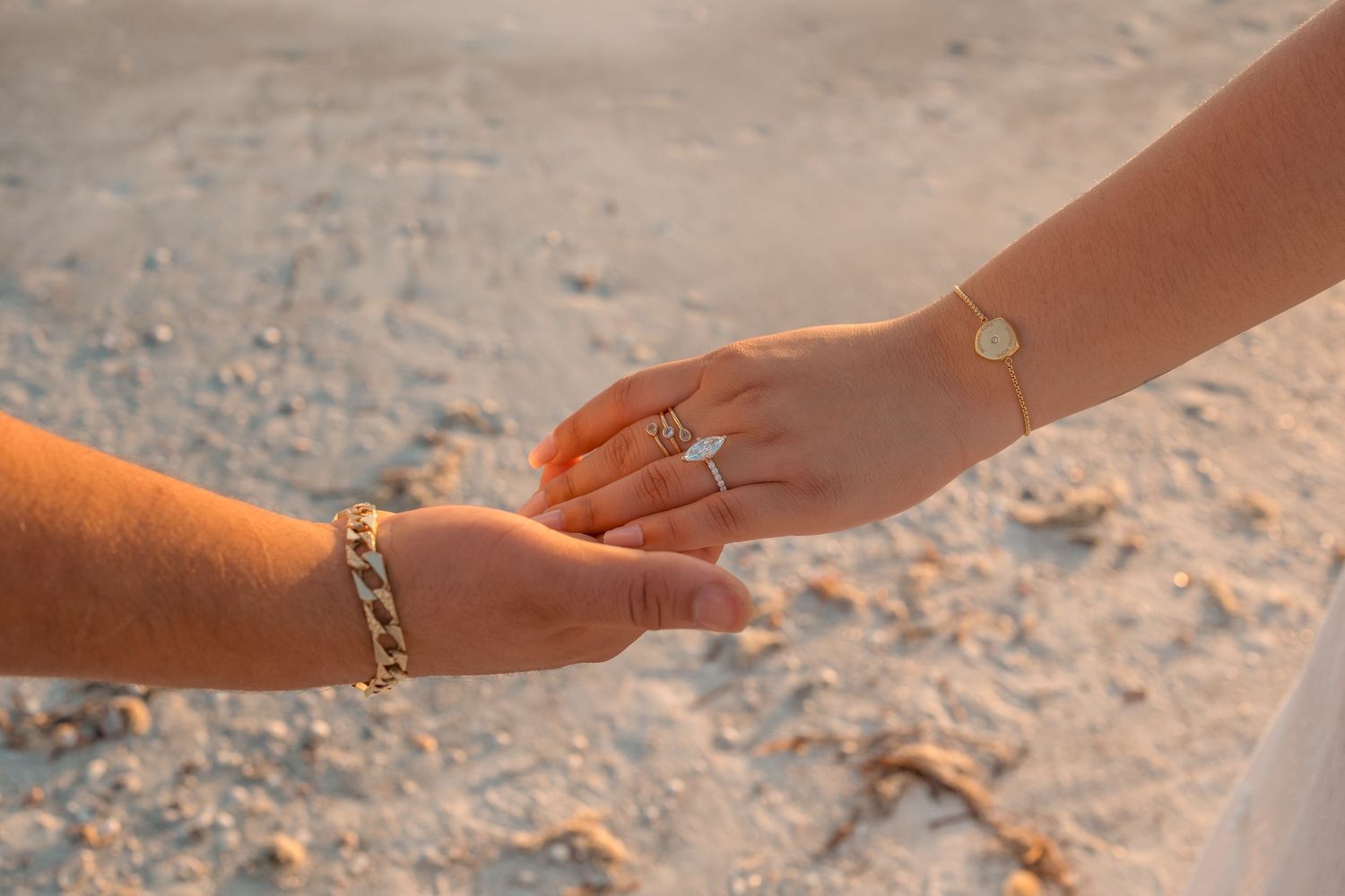 Two hands touching on a sandy beach, one wearing a diamond engagement ring and gold bracelet.