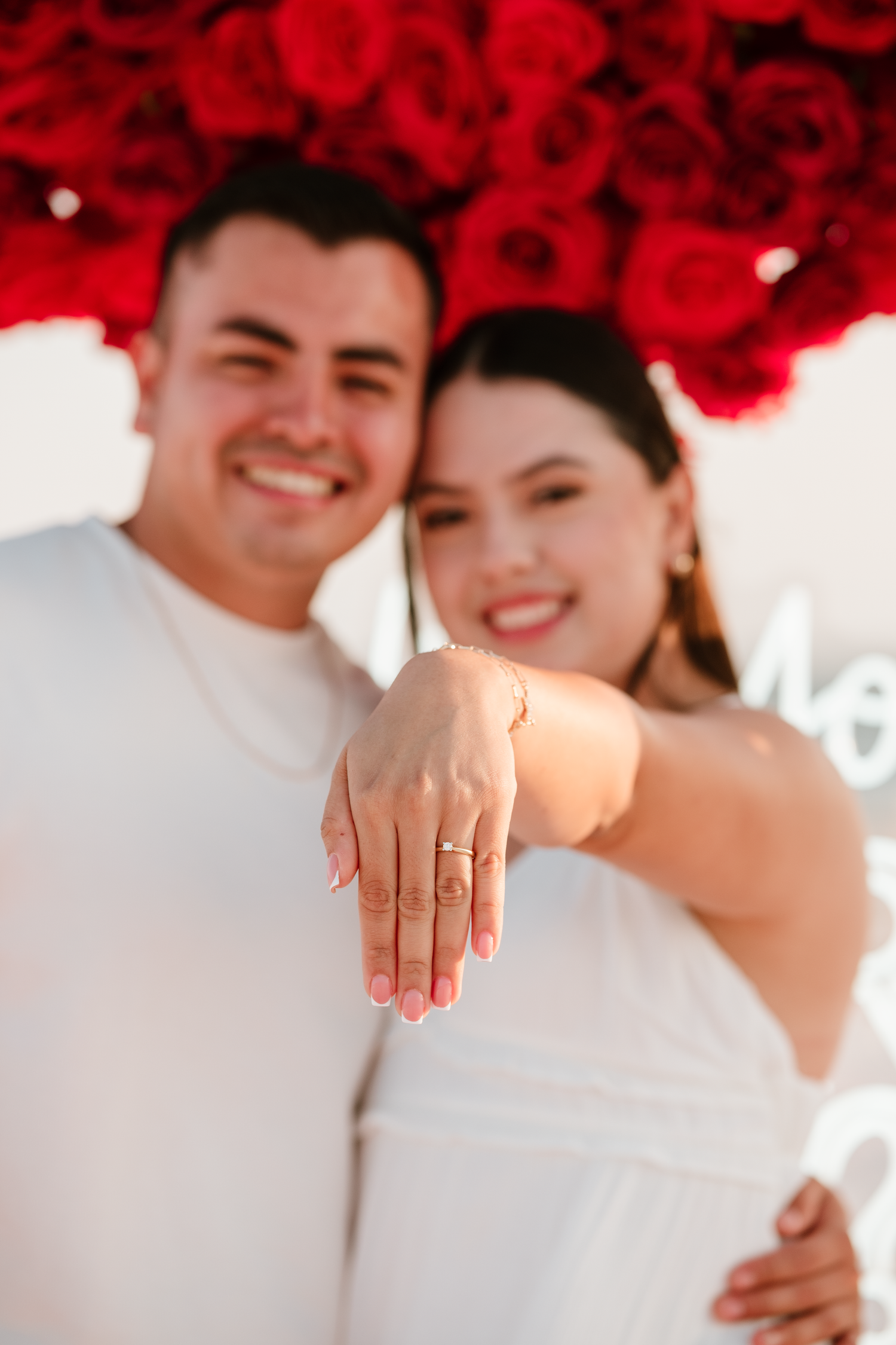 Happy couple in white outfits showing off engagement ring with red roses backdrop.
