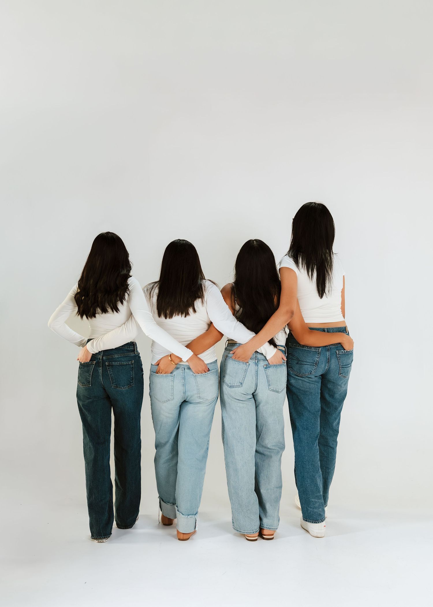 Four women with dark hair wearing white tops and jeans pose with backs to camera in a studio setting.