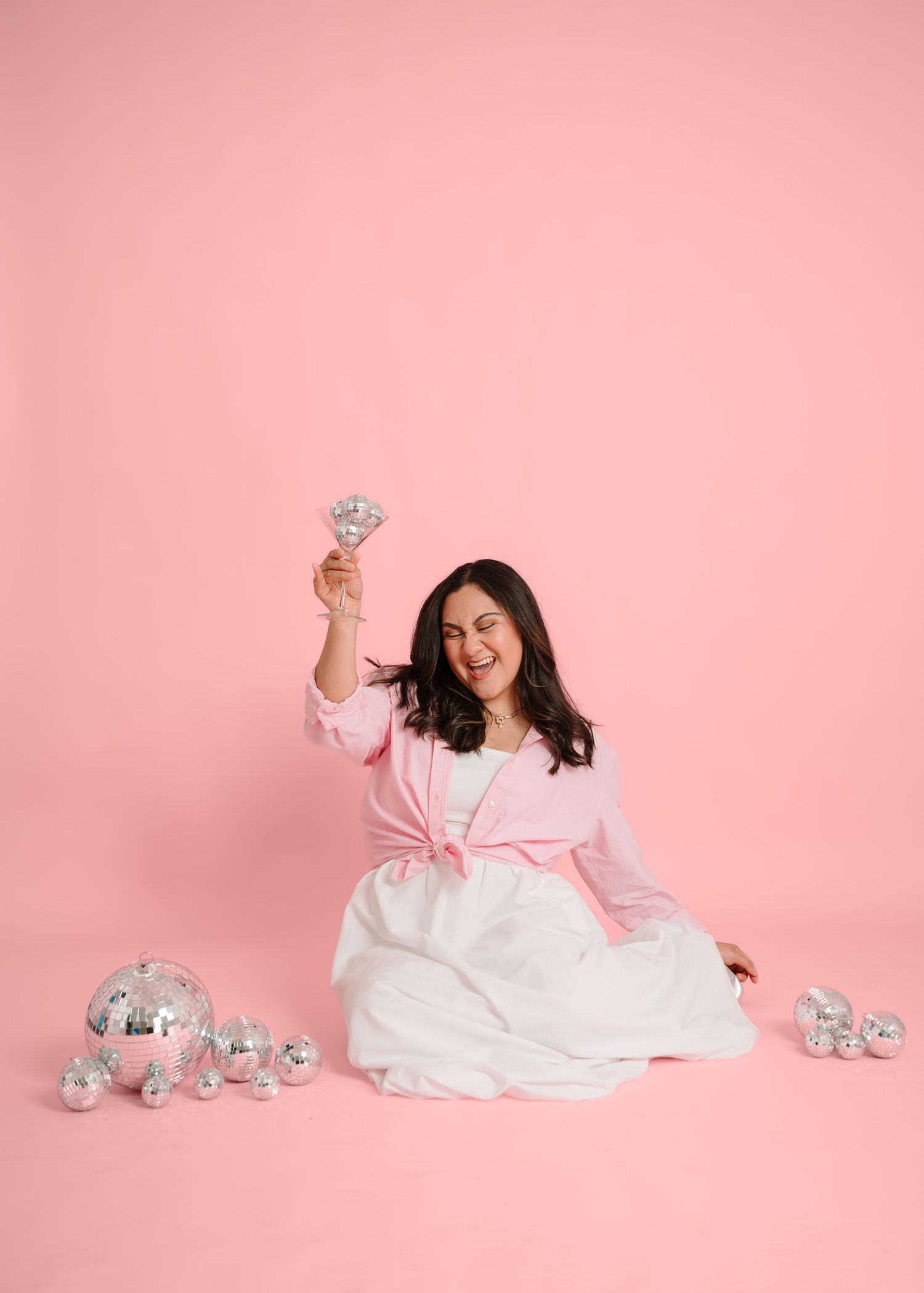 Woman in pink cardigan and white dress sits on pink backdrop holding a disco ball, surrounded by silver mirror balls.