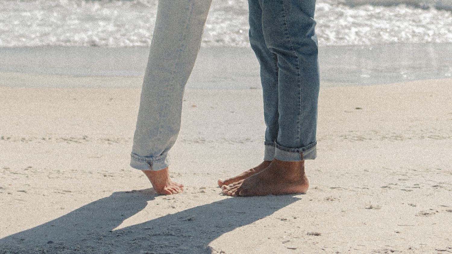 Two people standing barefoot on sandy beach in rolled-up jeans, waves glistening in background.