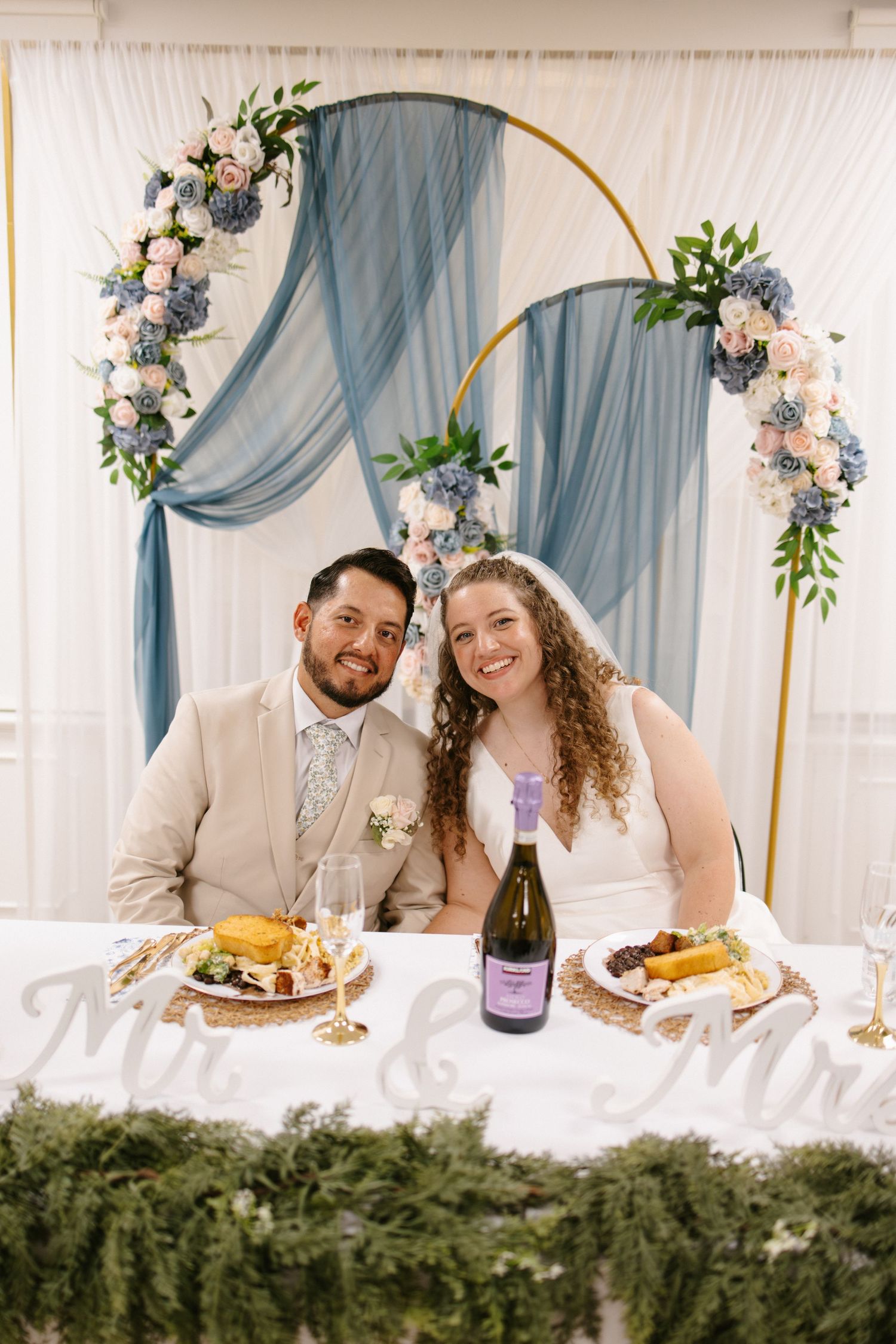 Newlyweds smile at sweetheart table with floral arch, champagne, and Mr. & Mrs. sign at wedding reception.