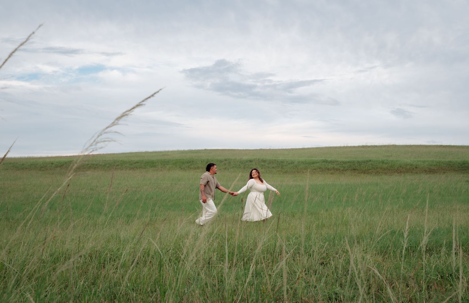 A person walks through a vast green prairie field with tall grass swaying in the wind on a cloudy day.