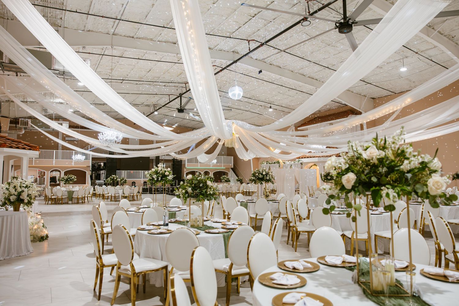 Spacious wedding reception with white draping, gold chairs, and tall floral centerpieces.