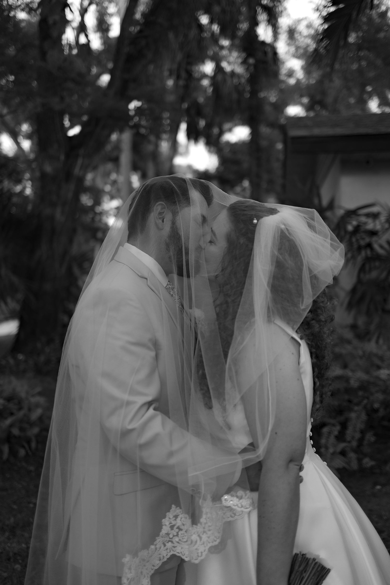 Black and white photo of bride and groom kissing under wedding veil outdoors among lush trees.