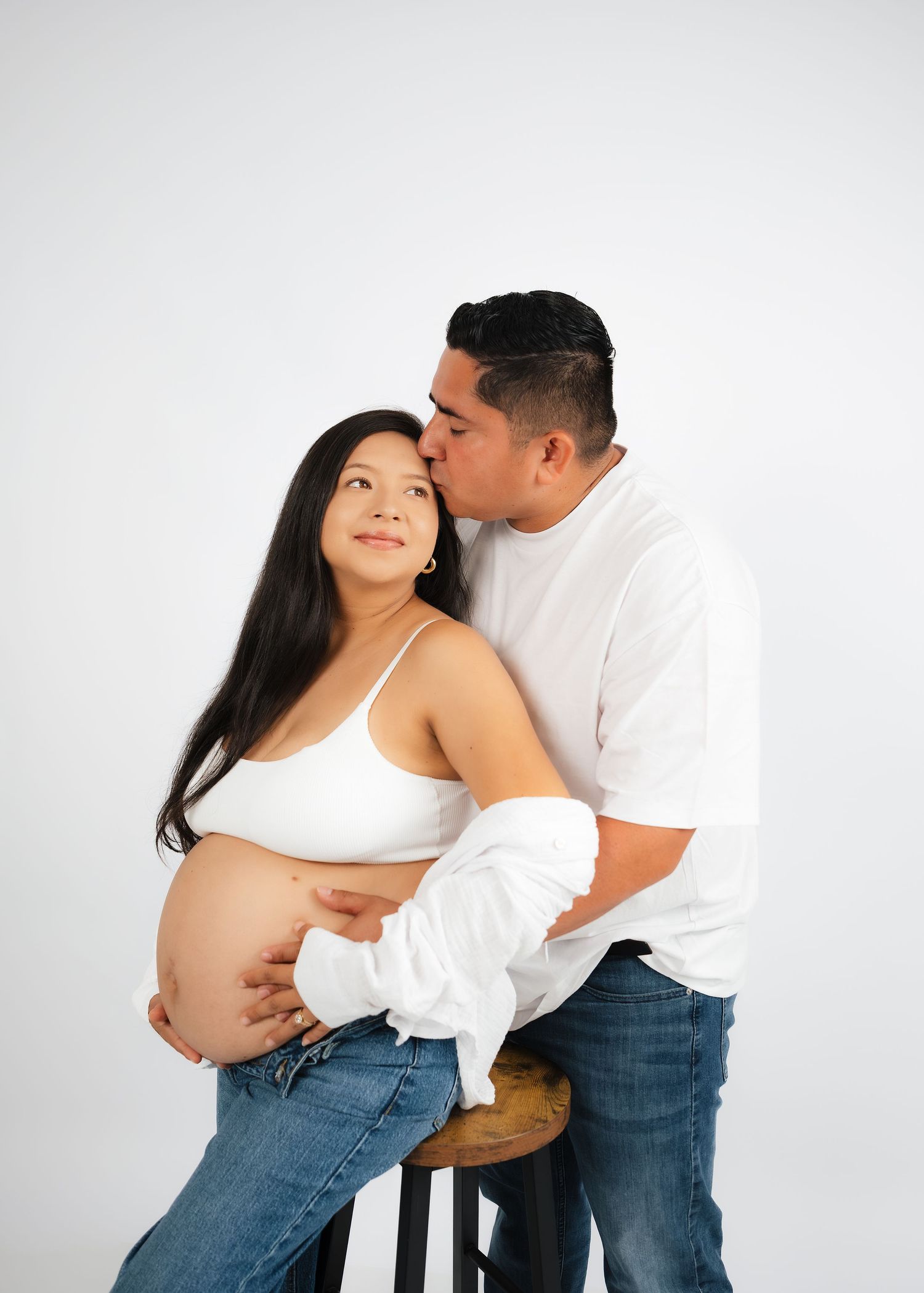 Pregnant woman in white crop top and jeans sits on stool as partner in white shirt kisses her cheek in studio.