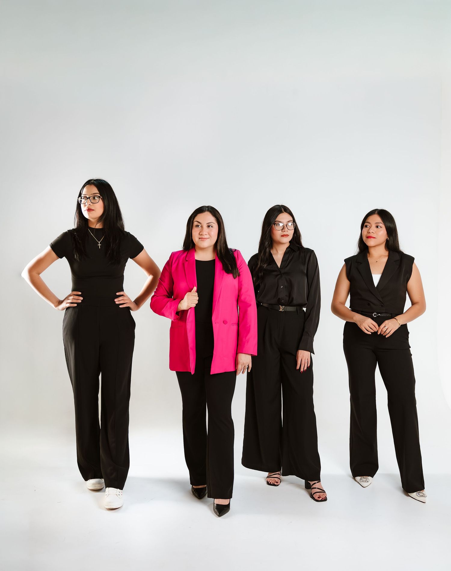 Four professional women in black outfits, one in a pink blazer, posing confidently in a studio setting.