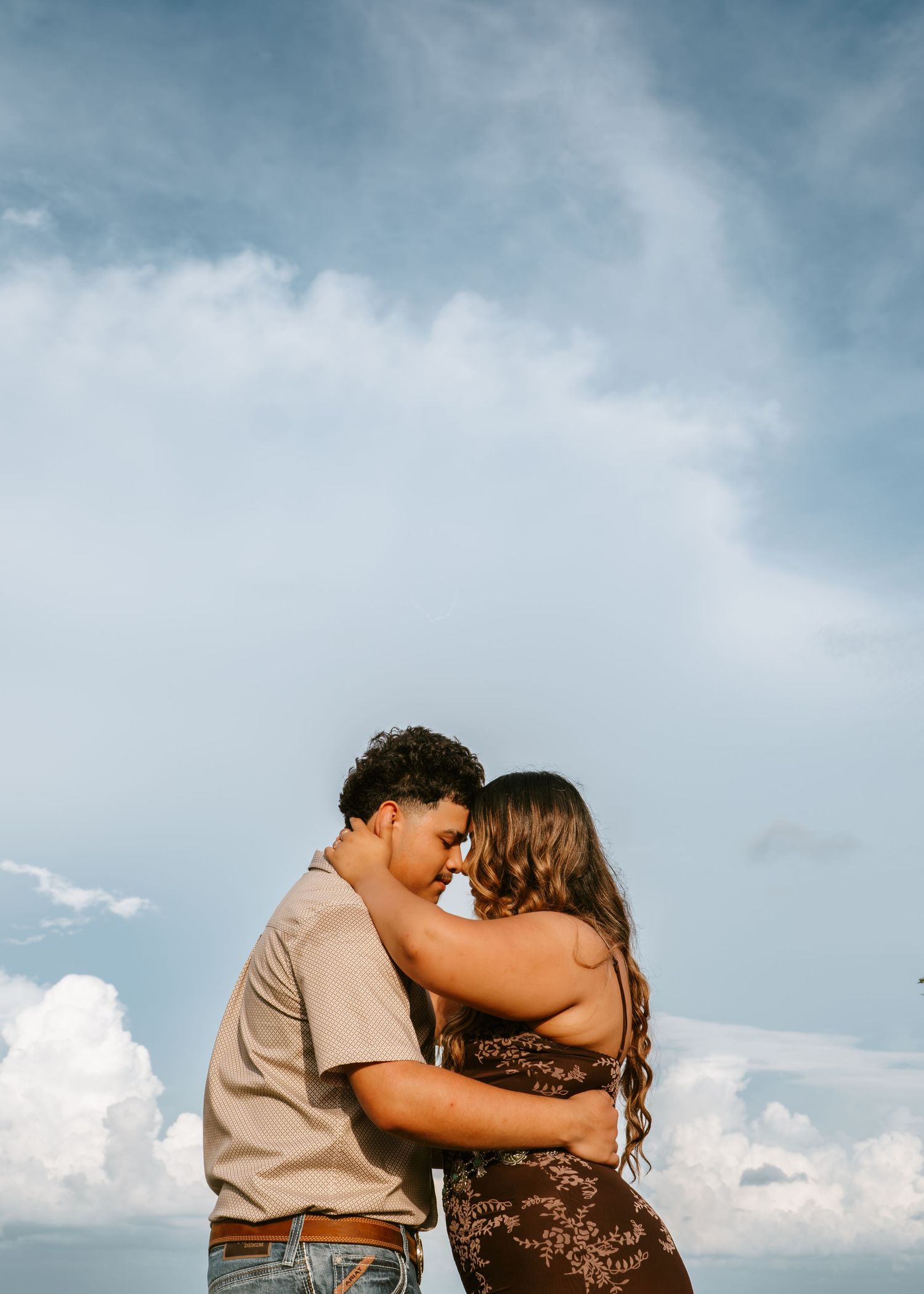 Romantic couple embracing and touching foreheads under a dramatic blue sky with fluffy white clouds.