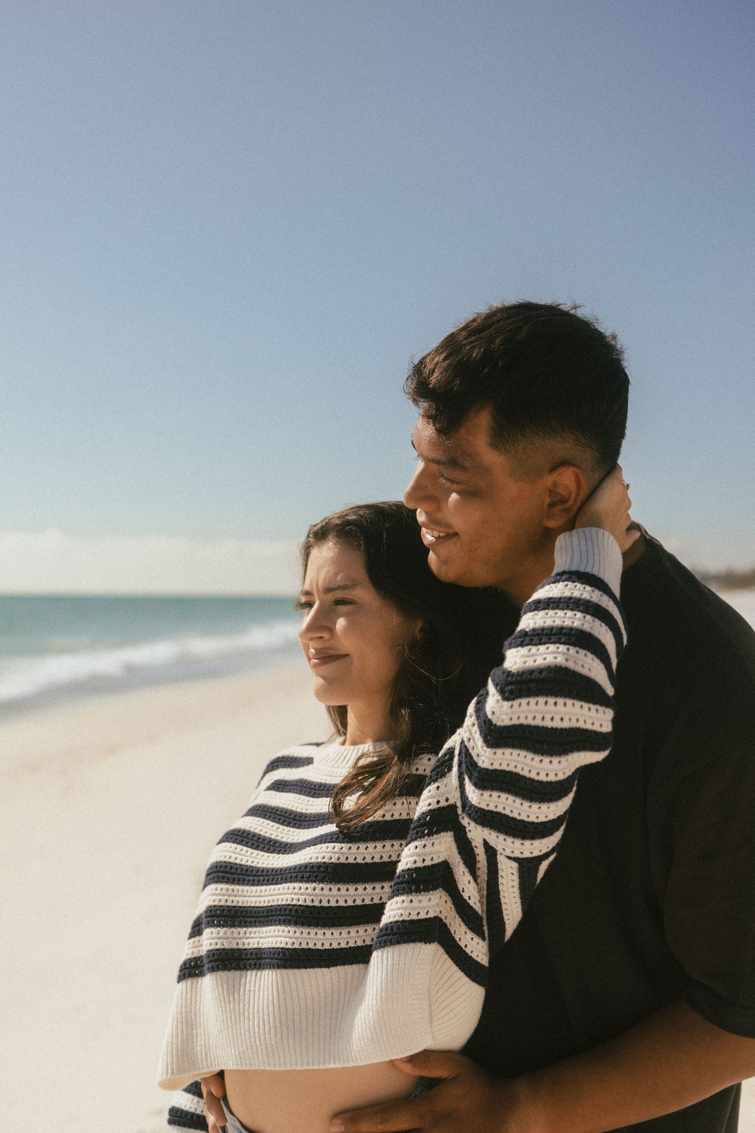 A young couple embraces on a sunny beach, smiling together with ocean waves and blue sky in the background.