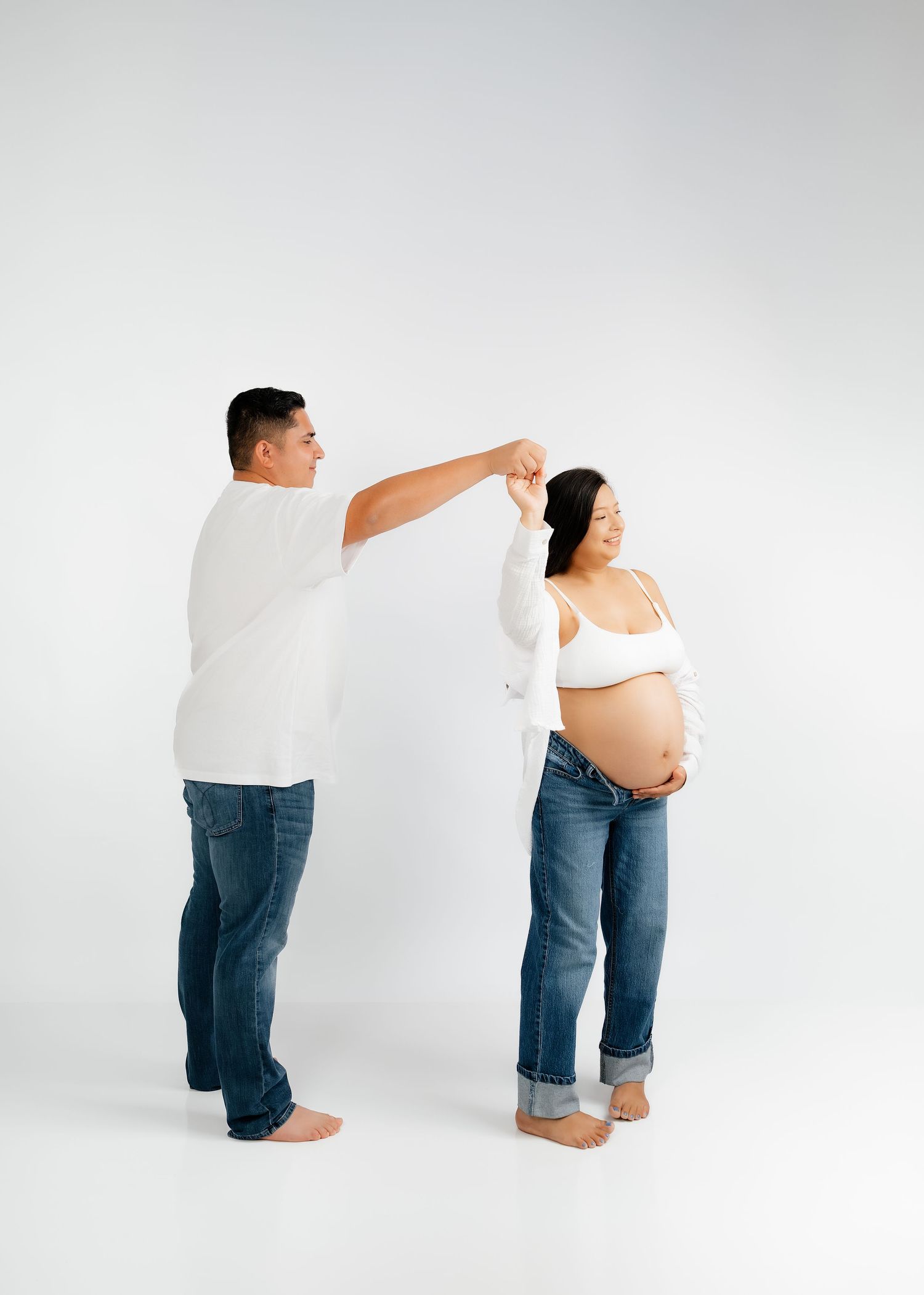 A man twirls a smiling pregnant woman in jeans and white top against a clean white studio background.