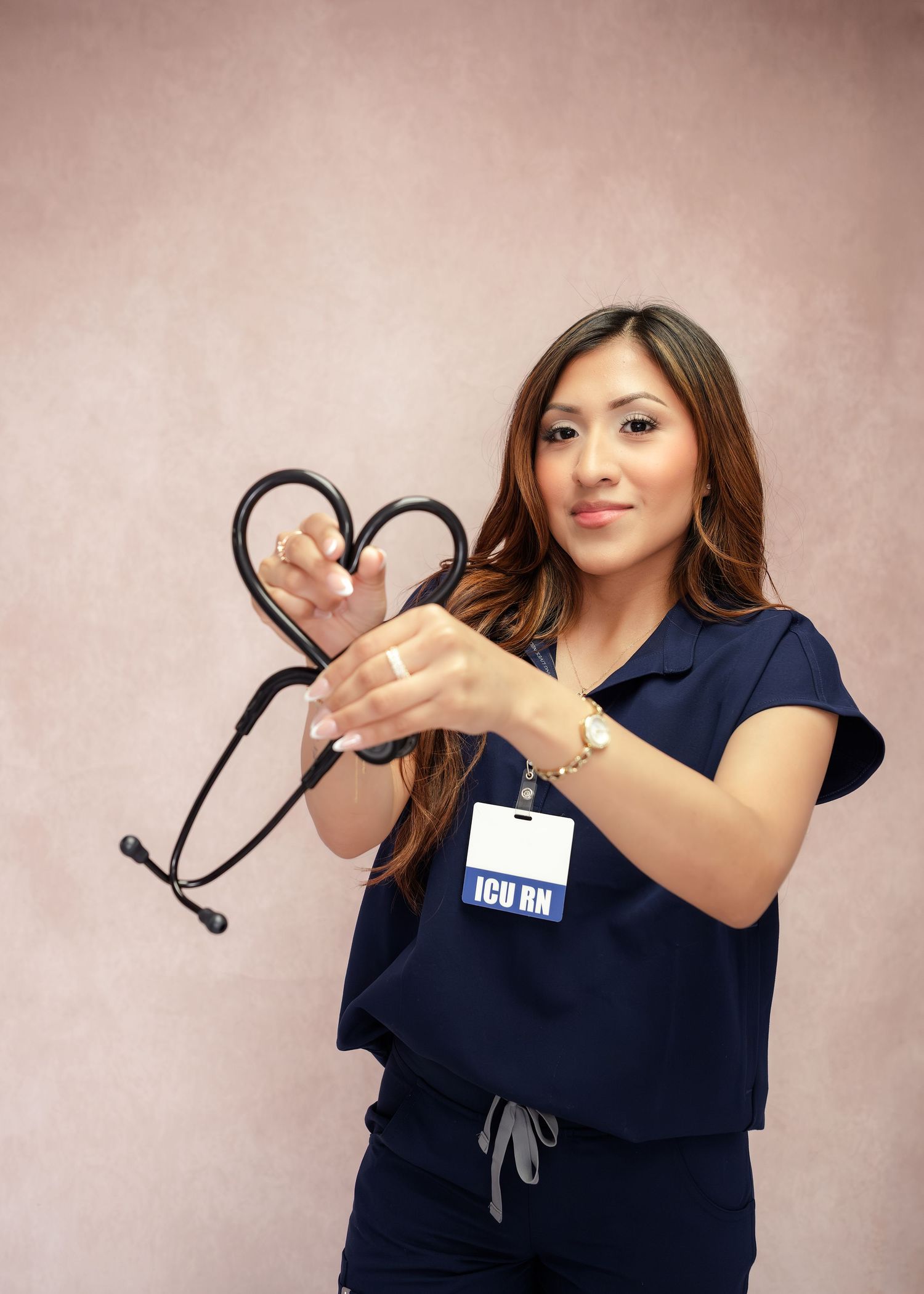 ICU RN nurse in blue scrubs holding a stethoscope shaped into a heart, smiling warmly against a pink background.